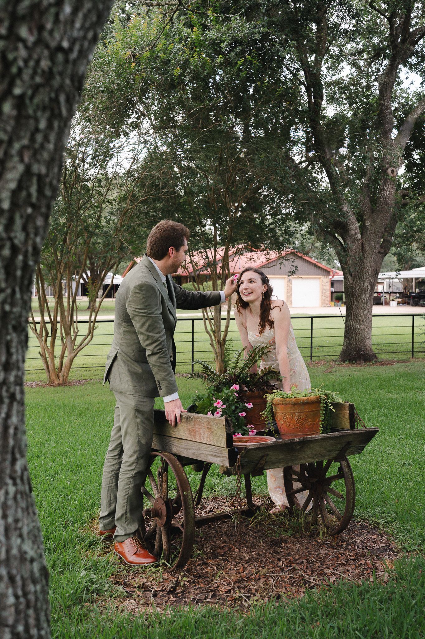 Bride and groom share a romantic moment beside a rustic wooden cart with flowers in a lush green outdoor setting.