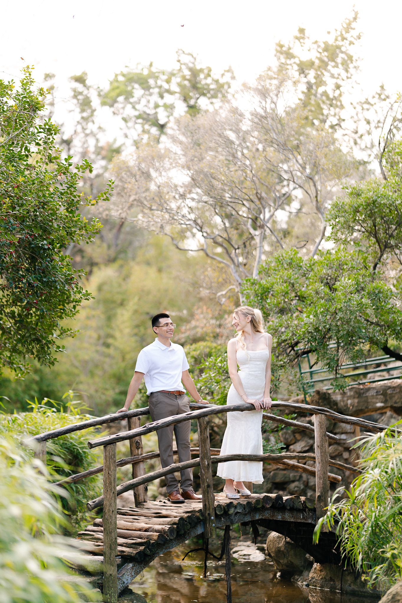 A couple sits together on a rustic wooden footbridge surrounded by lush greenery in a natural outdoor setting.
