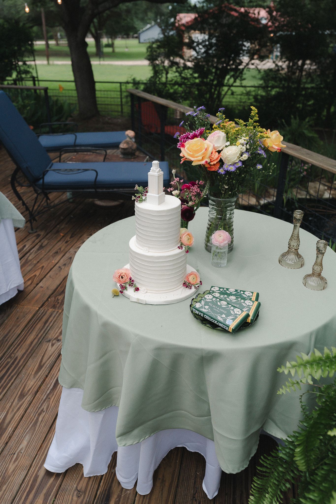 Elegant three-tier white wedding cake on sage green tablecloth with colorful floral arrangement on outdoor deck.