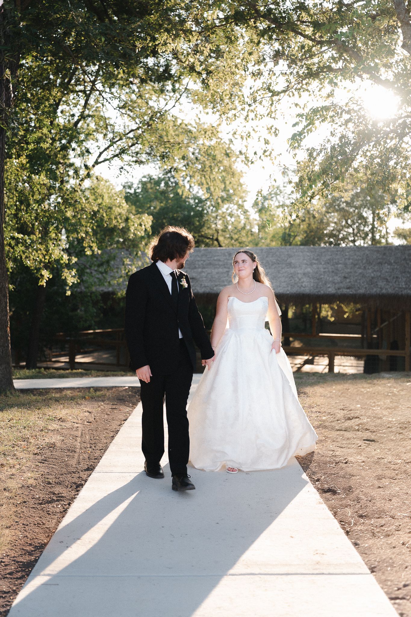 Bride and groom walk hand-in-hand down a sunlit pathway surrounded by lush trees at an outdoor wedding venue.