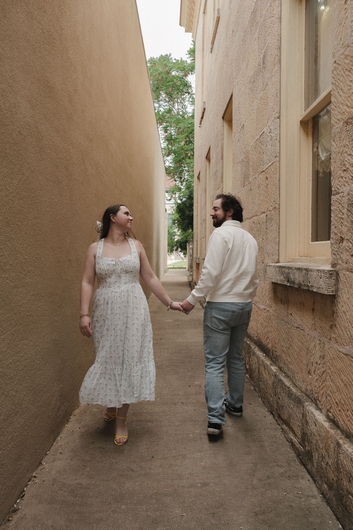 Couple holding hands walking through a narrow alley between stone buildings, woman in white floral dress, man in casual attire.
