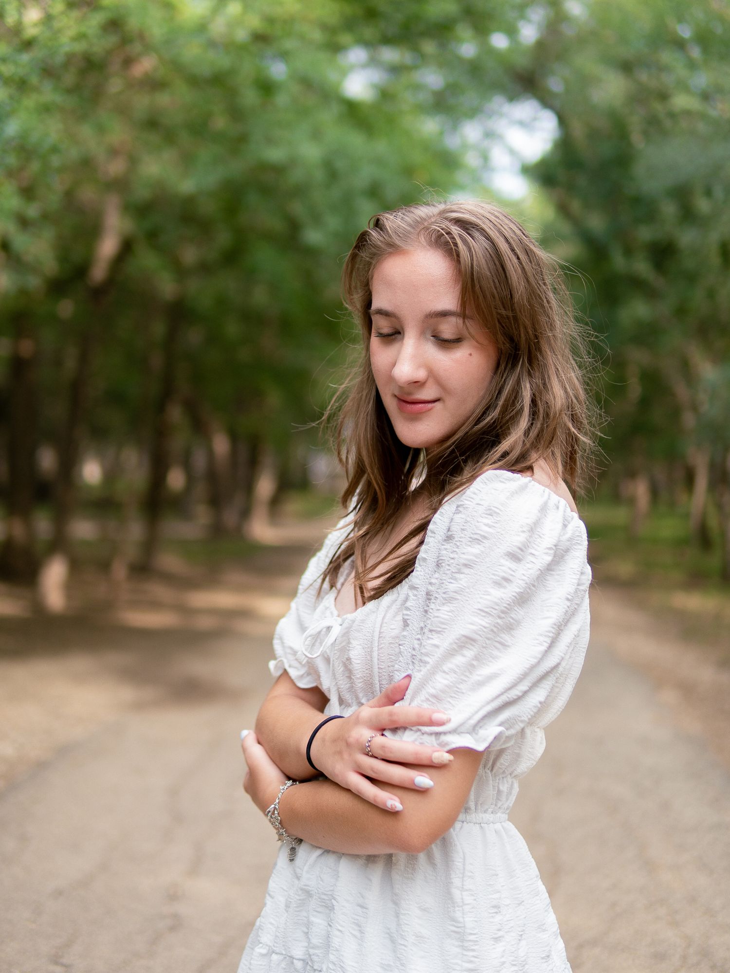 A person in a romantic white dress stands on a dirt path surrounded by green trees.