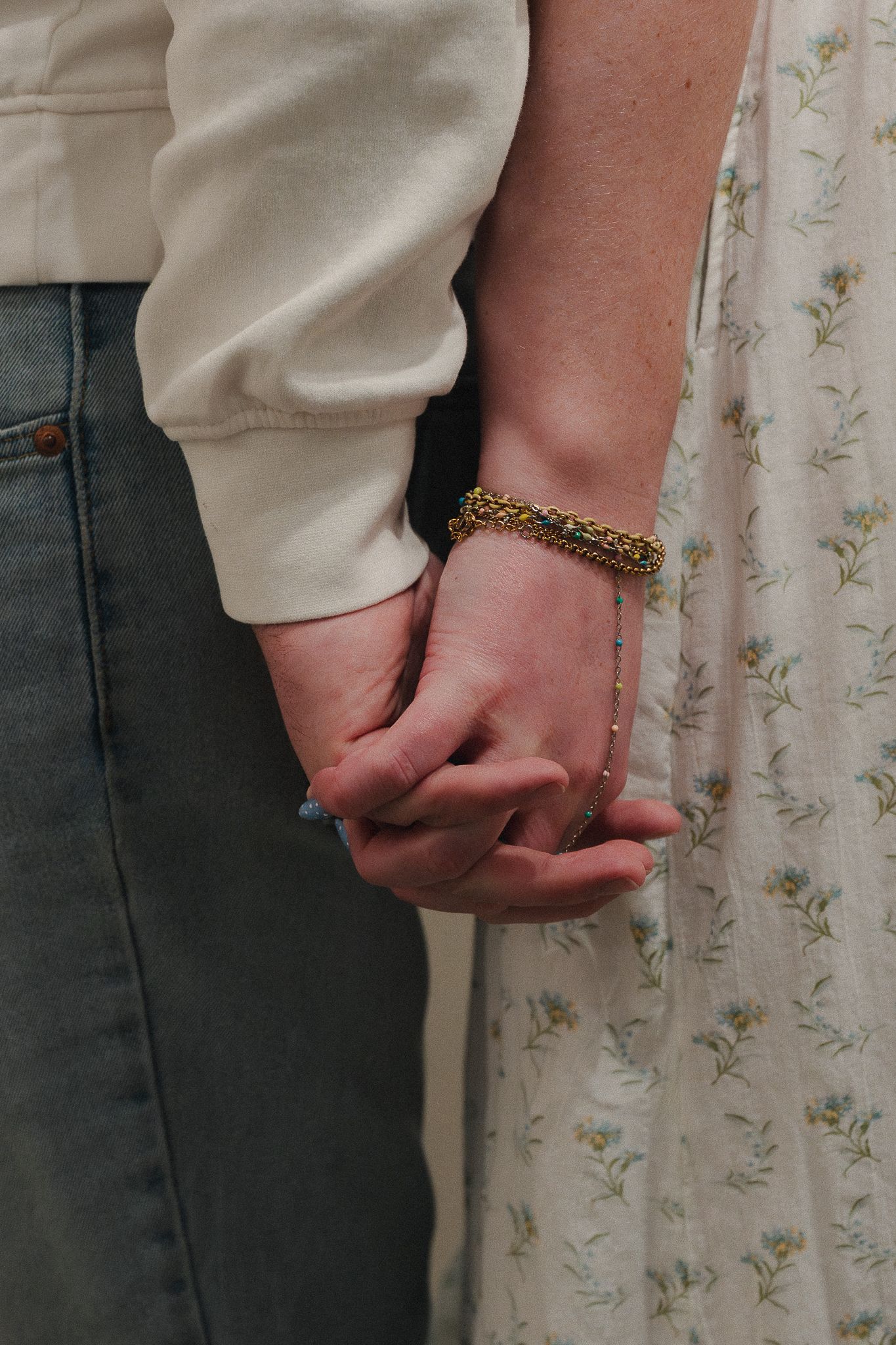Close-up of two people holding hands, one wearing a gold chain bracelet, floral dress and jeans visible.