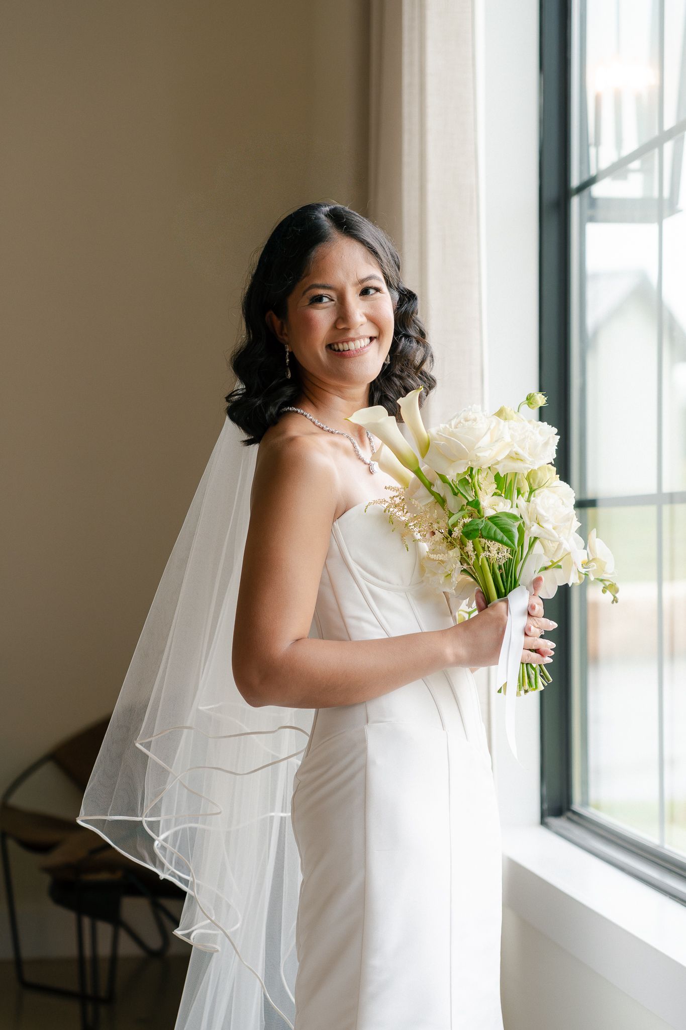 A bride in a white wedding dress holds a beautiful bouquet of white lilies and flowers while standing by a window.