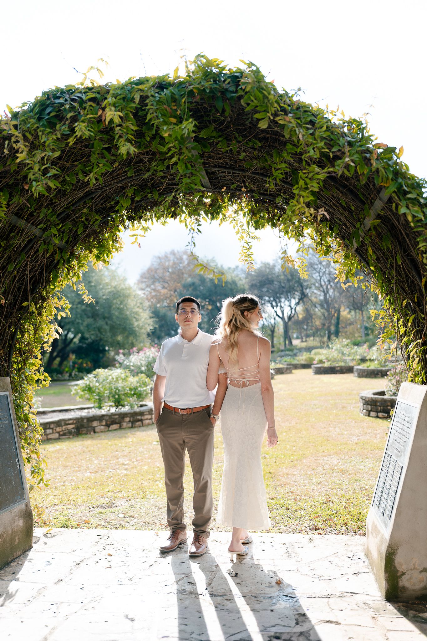 A couple stands beneath a lush green garden archway in a sunlit outdoor setting during a romantic photoshoot.