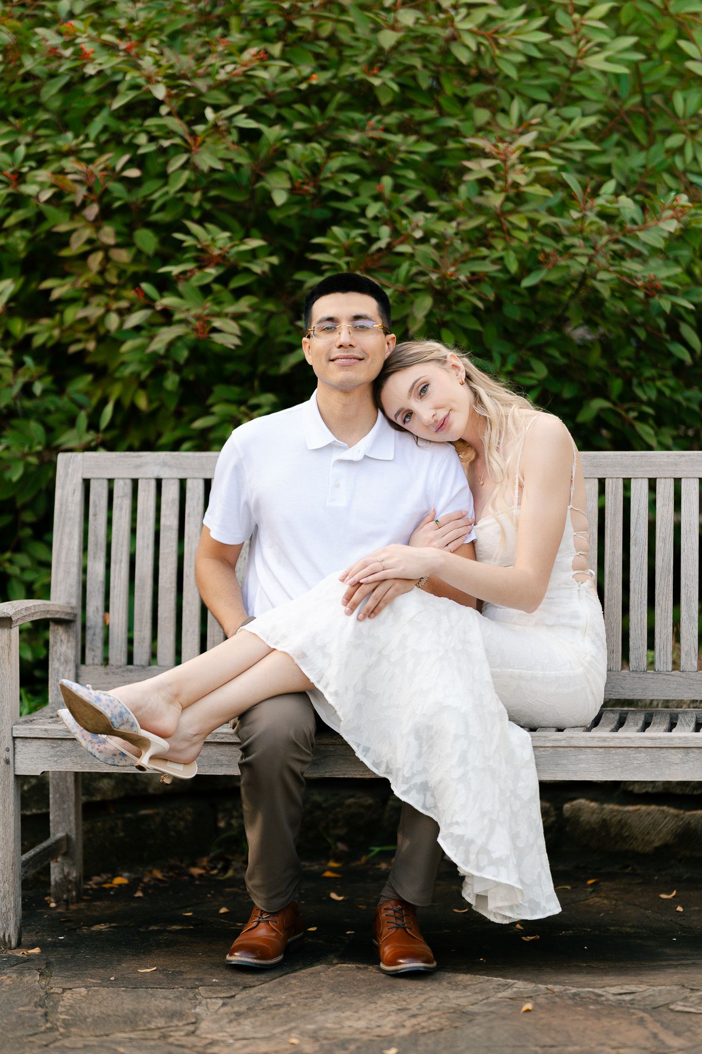 A couple sits together on a wooden garden bench, with the bride wearing a white dress and groom in casual attire.