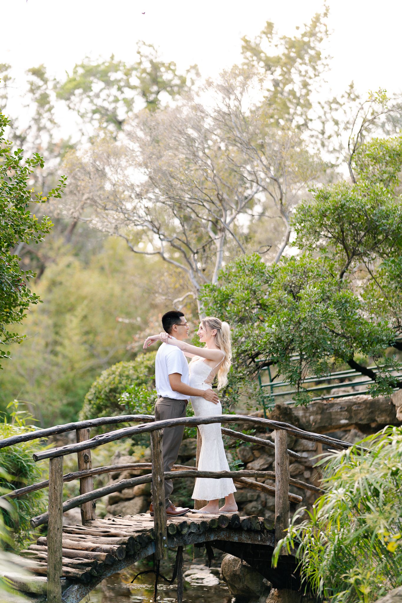 A couple shares a romantic moment on a rustic wooden bridge surrounded by lush greenery in a garden setting.