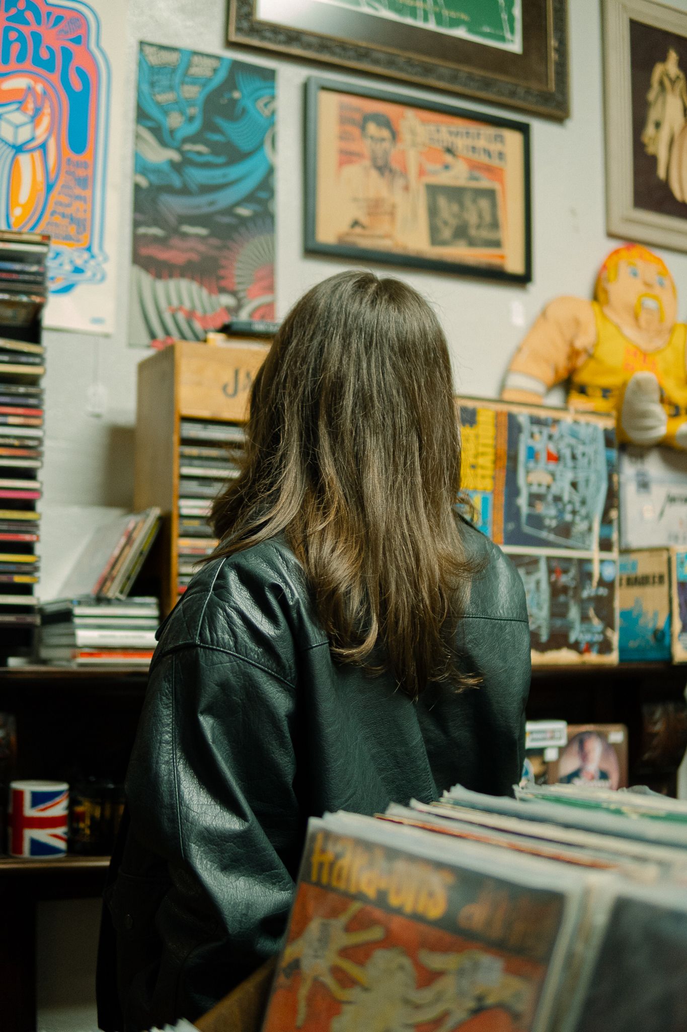 Someone in a black leather jacket browses through vinyl records in a colorful record store with art-covered walls.