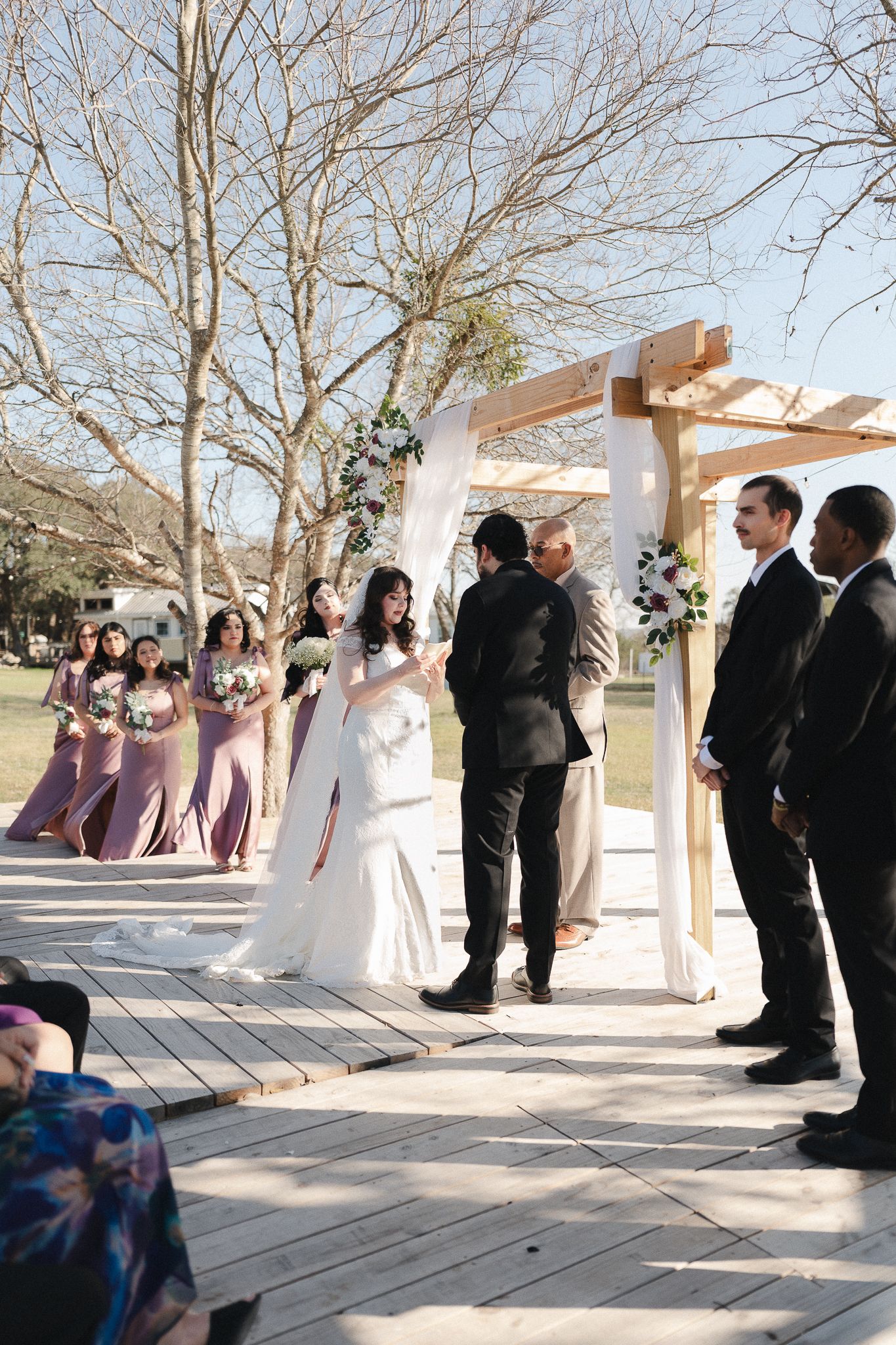 Bride and groom exchange vows under a wooden arch with bridesmaids in pink dresses at an outdoor wedding ceremony.
