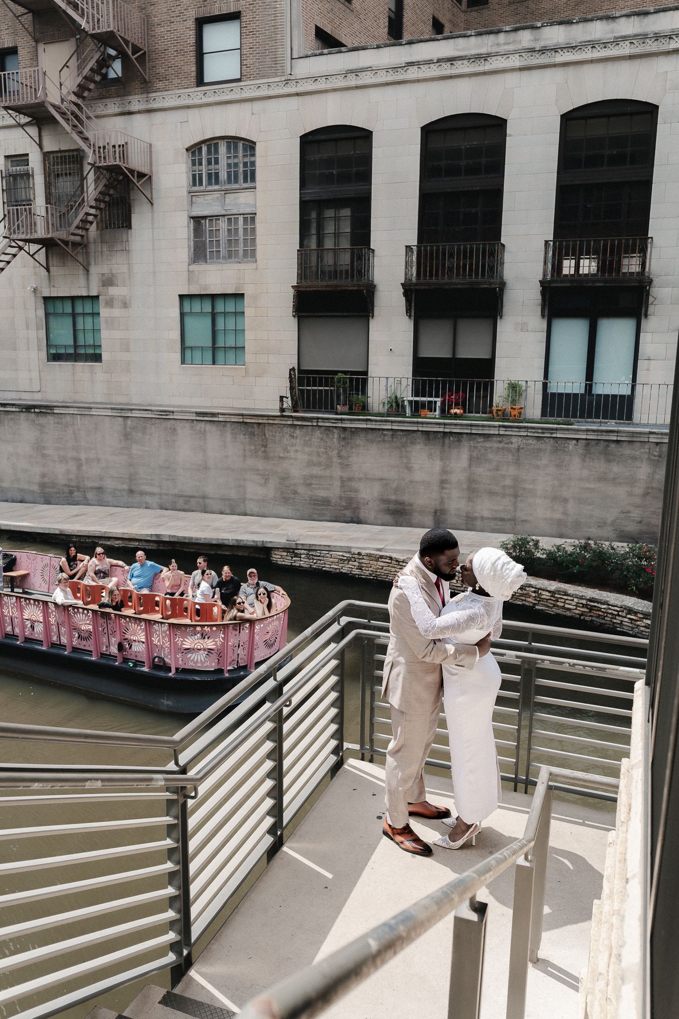 Couple in white attire embracing on a balcony overlooking the San Antonio River Walk with a tour boat passing by.