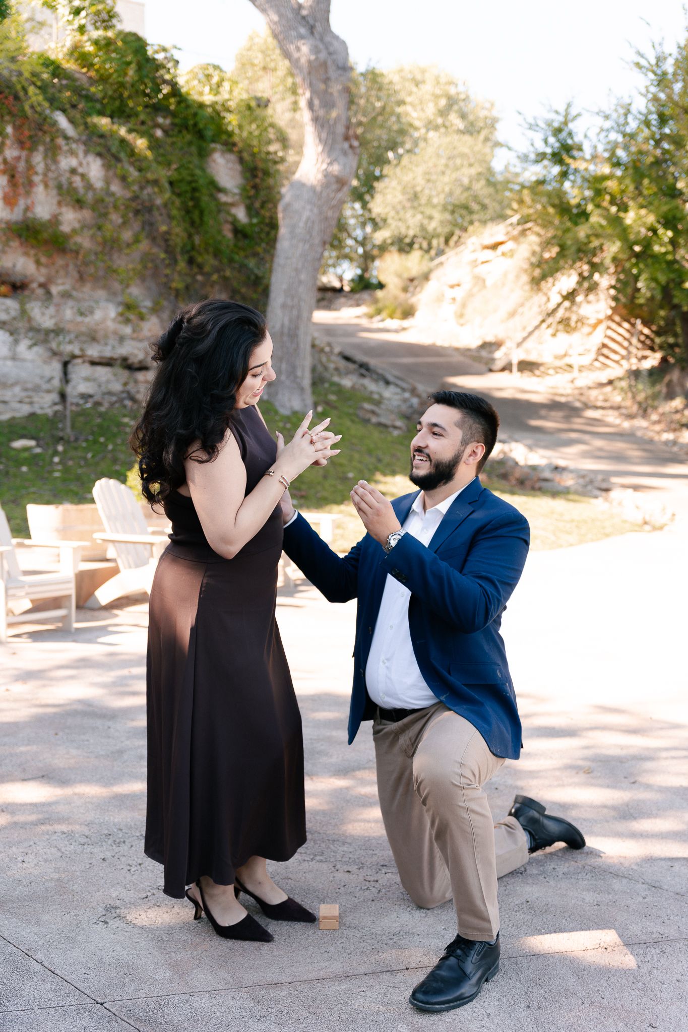 A couple in formal attire playfully interacts in a sunny outdoor setting with stone pathways and trees.