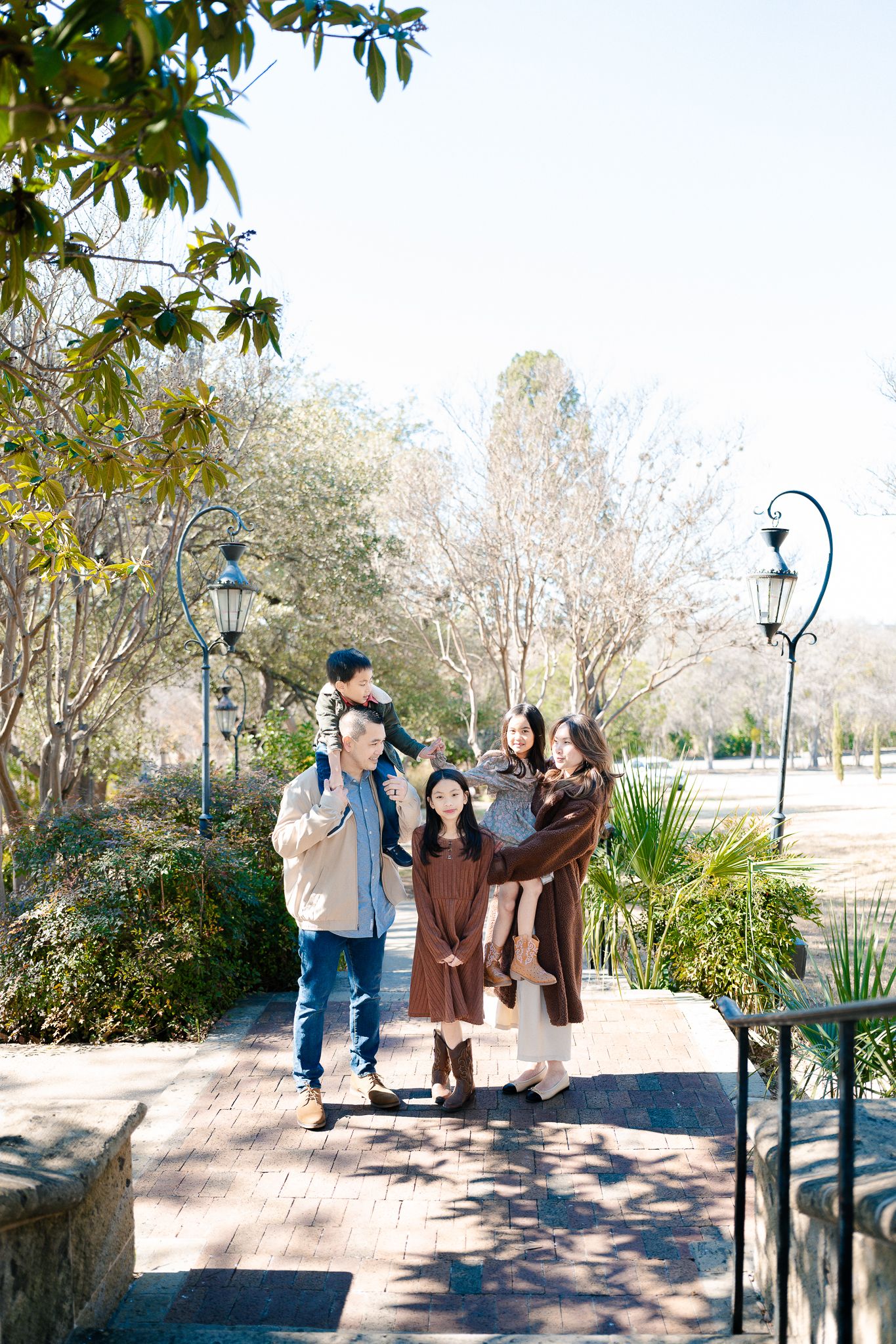 Family enjoying a sunny day together on a stone path with decorative lampposts and lush greenery in a park setting.
