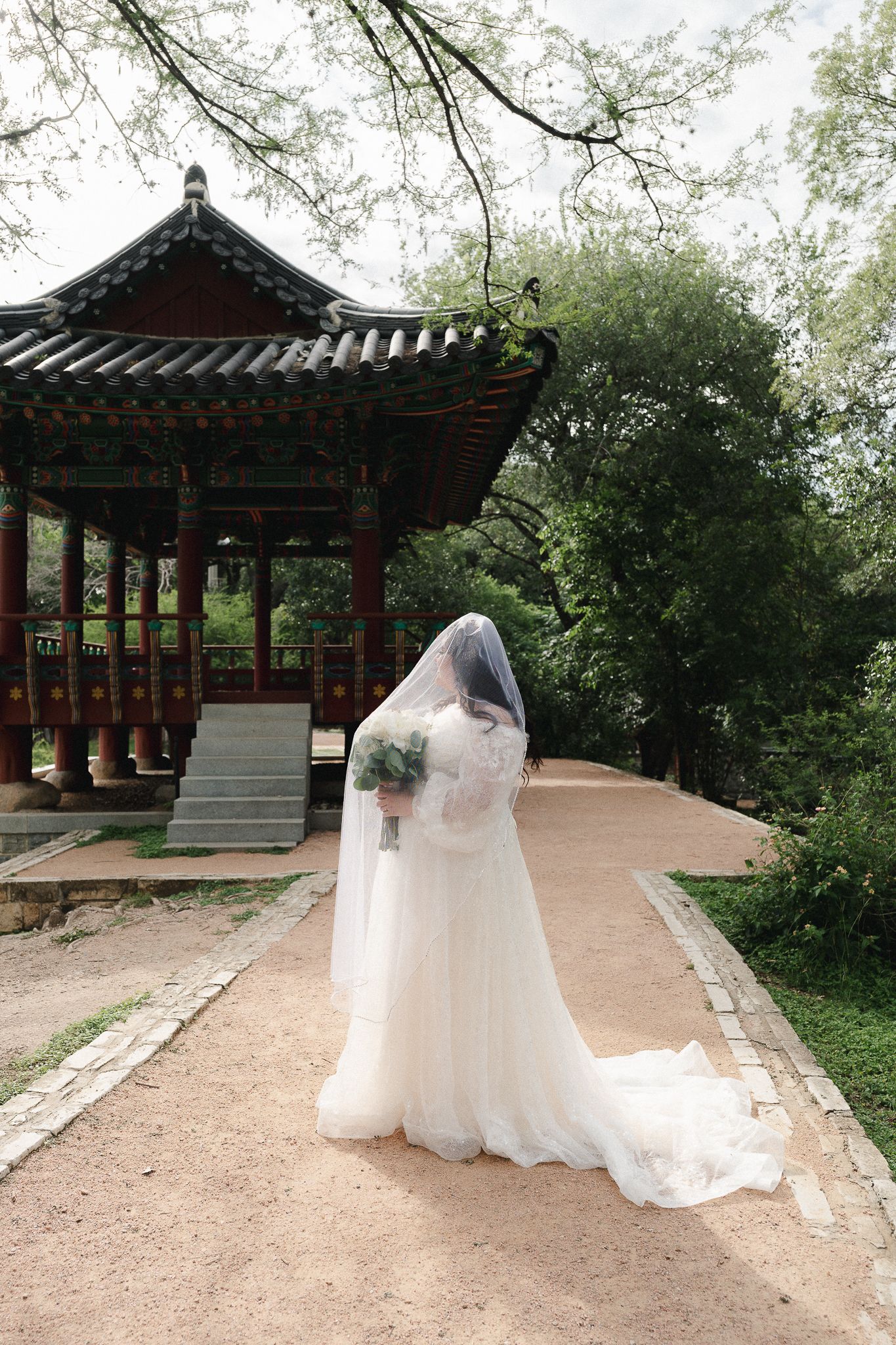 Bride in white gown and veil holds bouquet before a traditional Korean pavilion surrounded by lush greenery.