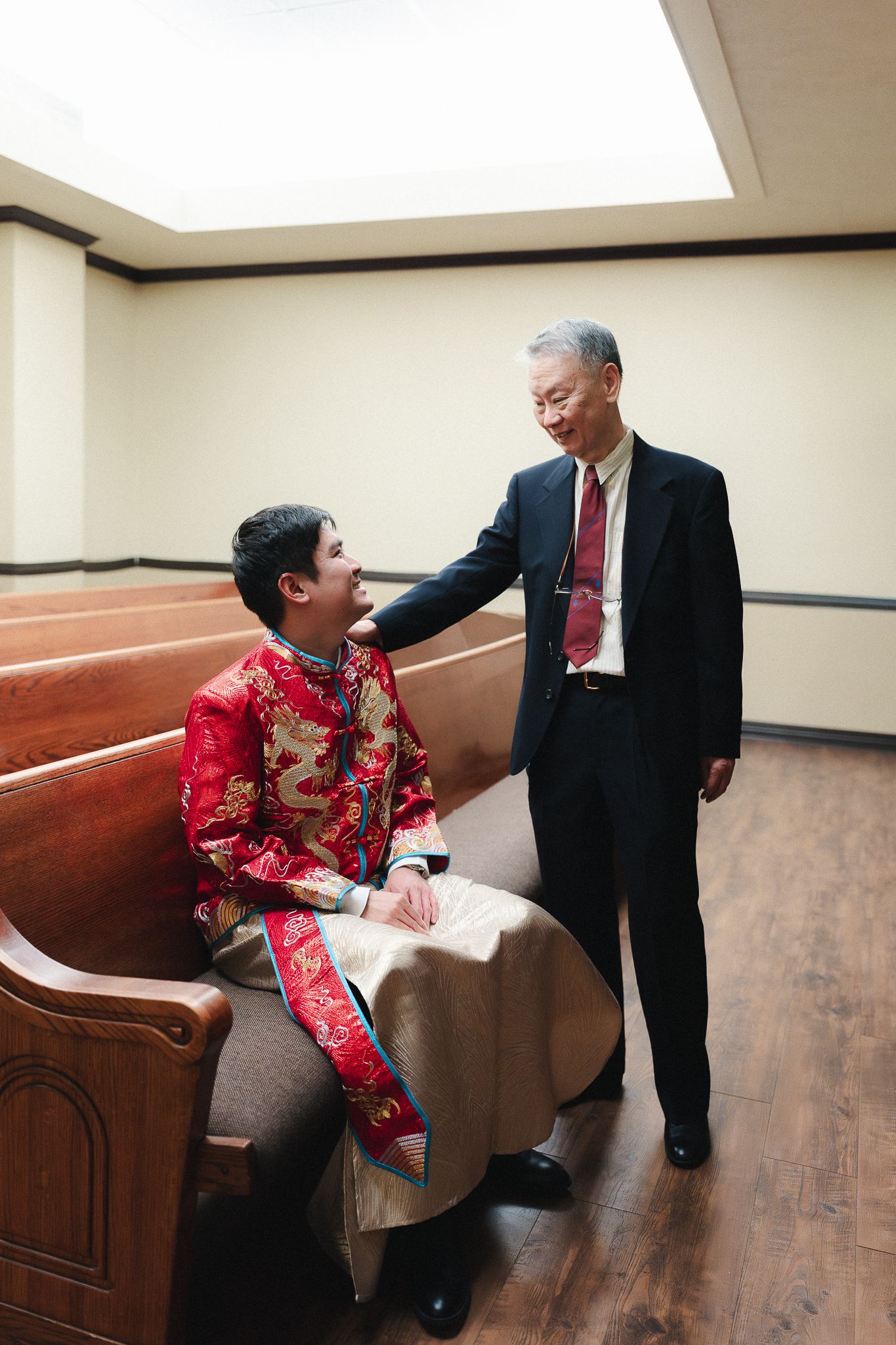 Two men in traditional Chinese and formal Western attire share a warm moment on a wooden church pew.