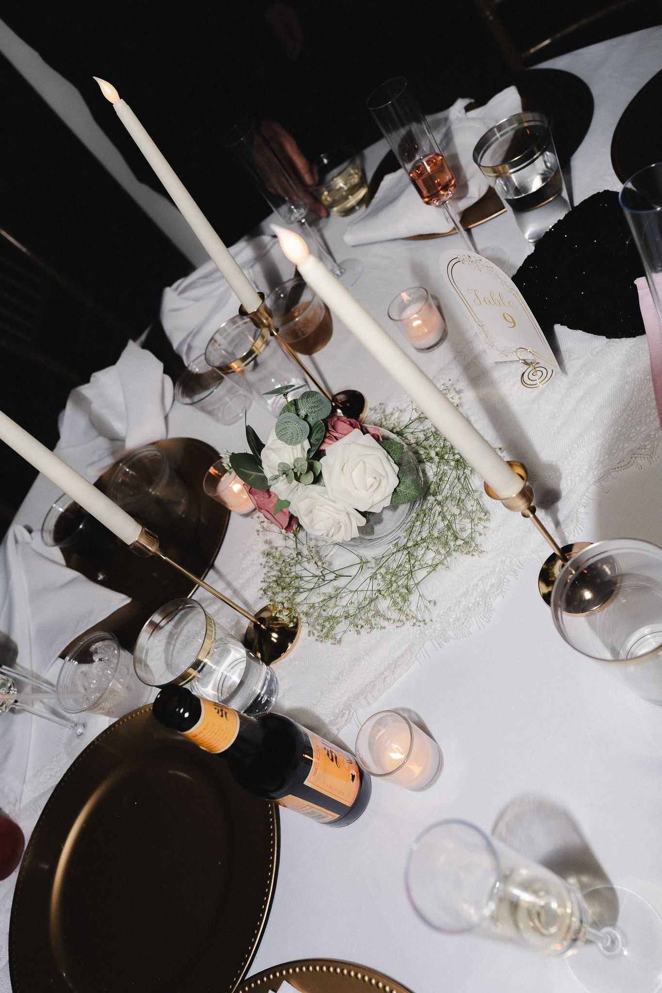 Elegant wedding table setting with white candles, gold plates, champagne bottles, floral centerpiece, and glowing candles on white linen.