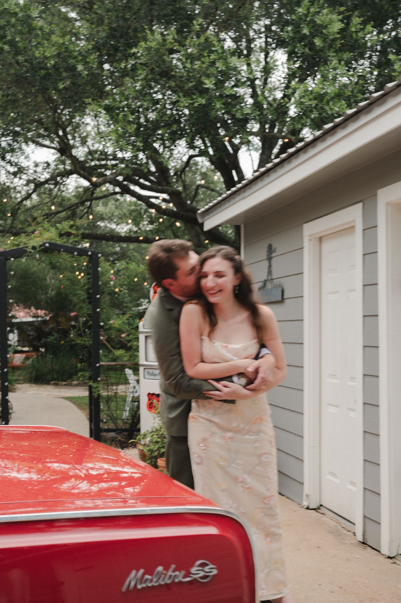 Couple embracing and kissing beside a classic red Chevrolet Malibu SS near a gray garage.