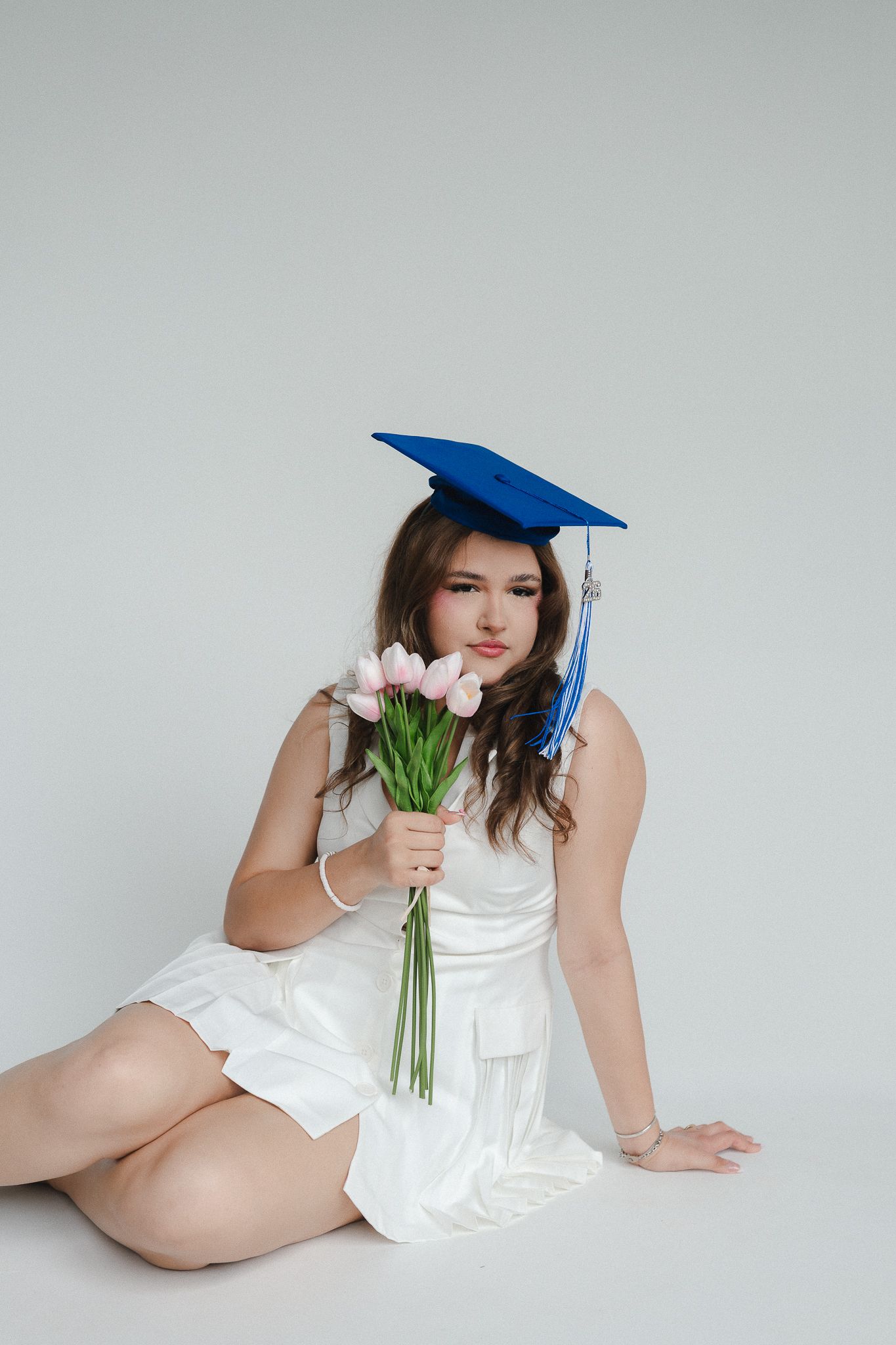 Young woman in white dress and blue graduation cap sitting on floor holding pink tulips bouquet.