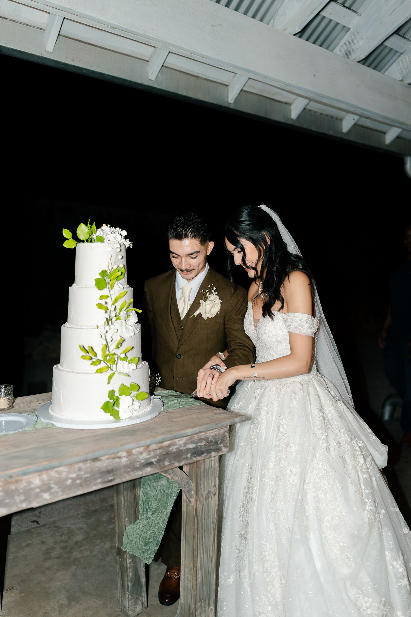 A couple cuts their elegant white wedding cake adorned with green foliage at their reception.