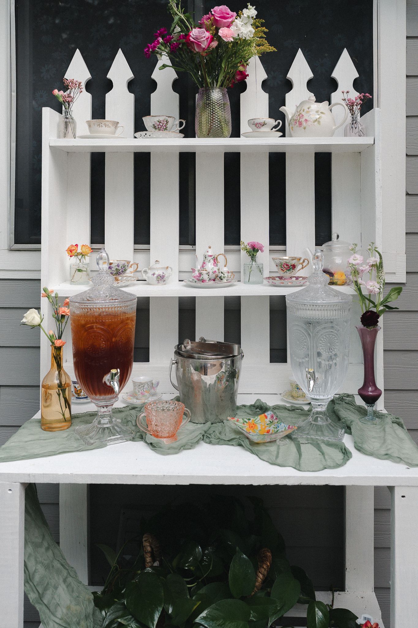 Vintage tea party display with picket fence shelves, floral arrangements, teacups, and beverage dispensers on a white table.