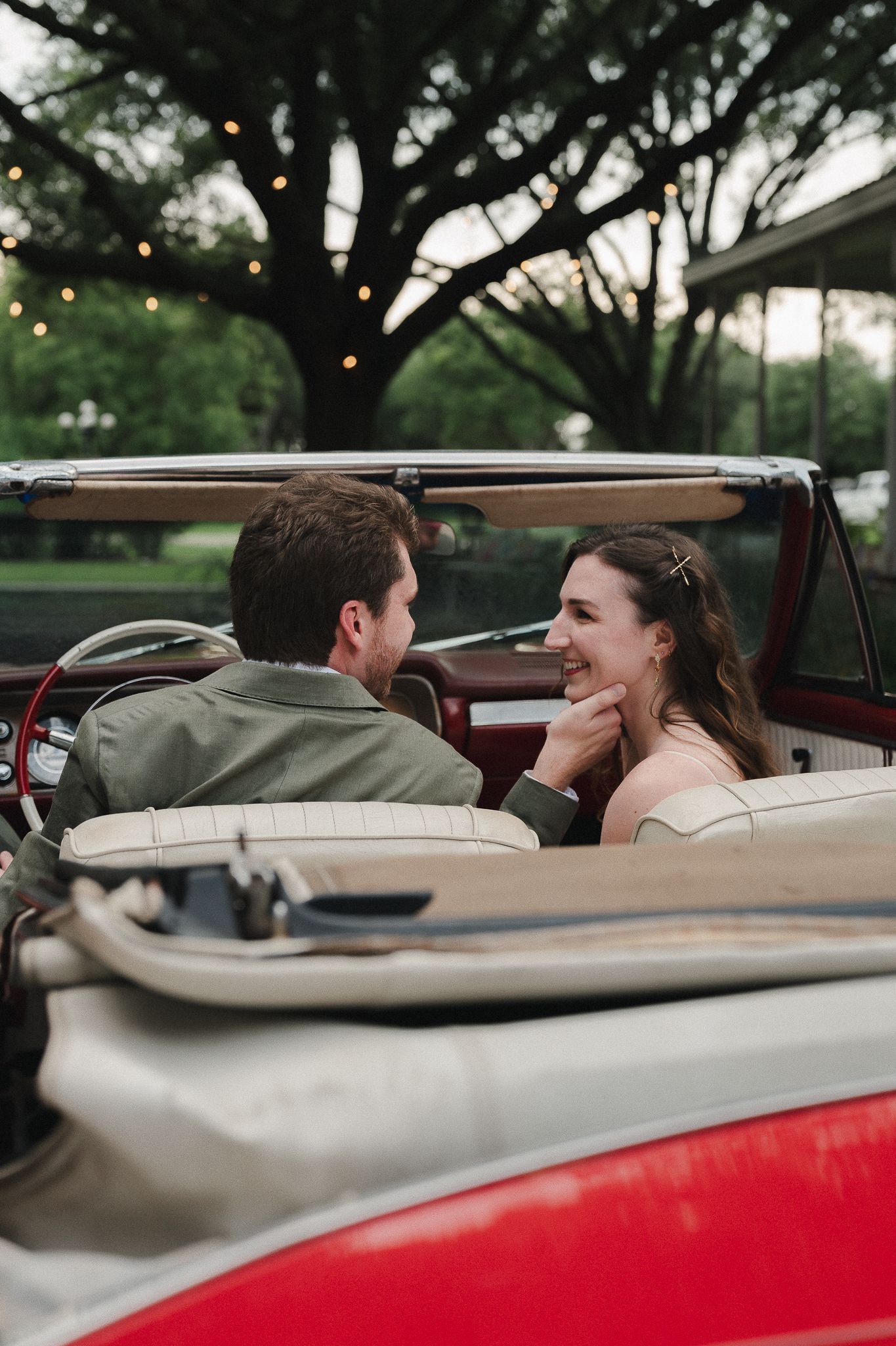 Couple sharing a romantic moment in a vintage red convertible car with string lights and trees in the background.