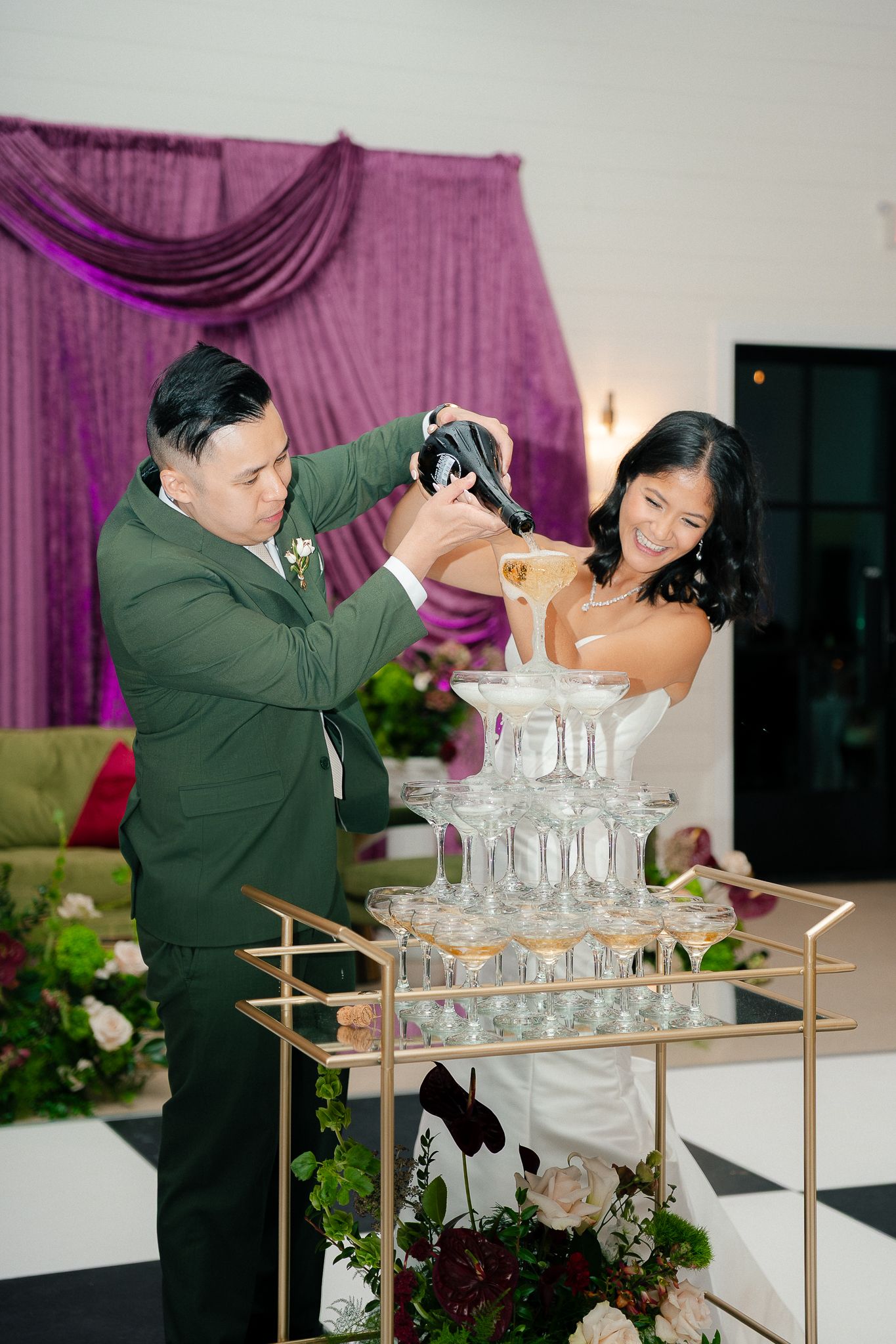 A person in a green jacket carefully pours drinks into a champagne tower at an elegant event with purple lighting.