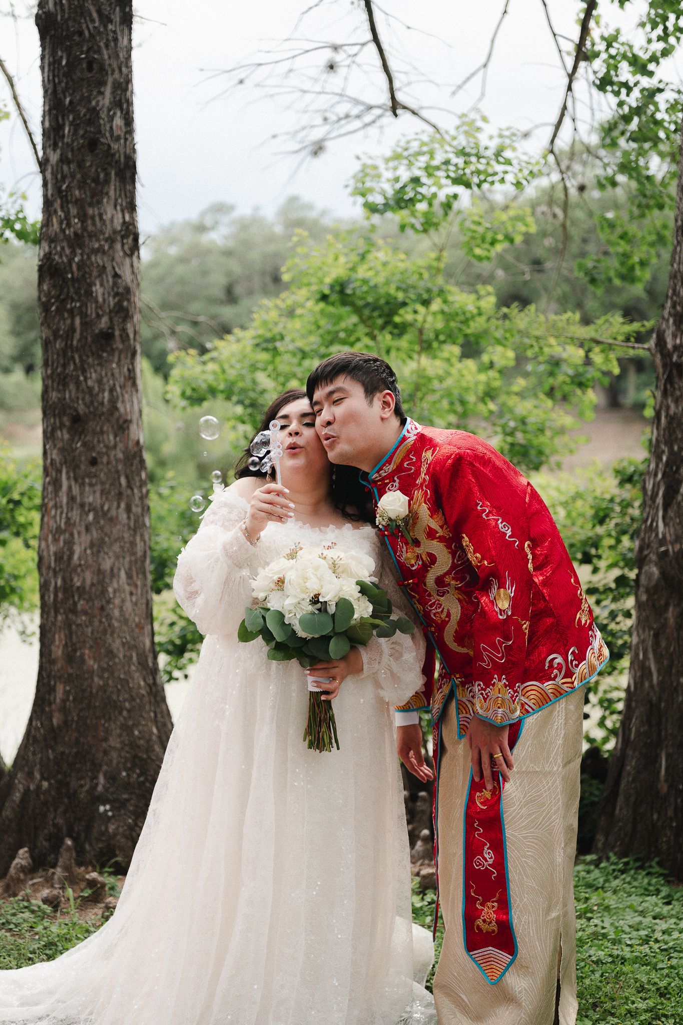 A bride in white lace blows bubbles while her groom in red Chinese traditional attire kisses her cheek outdoors.