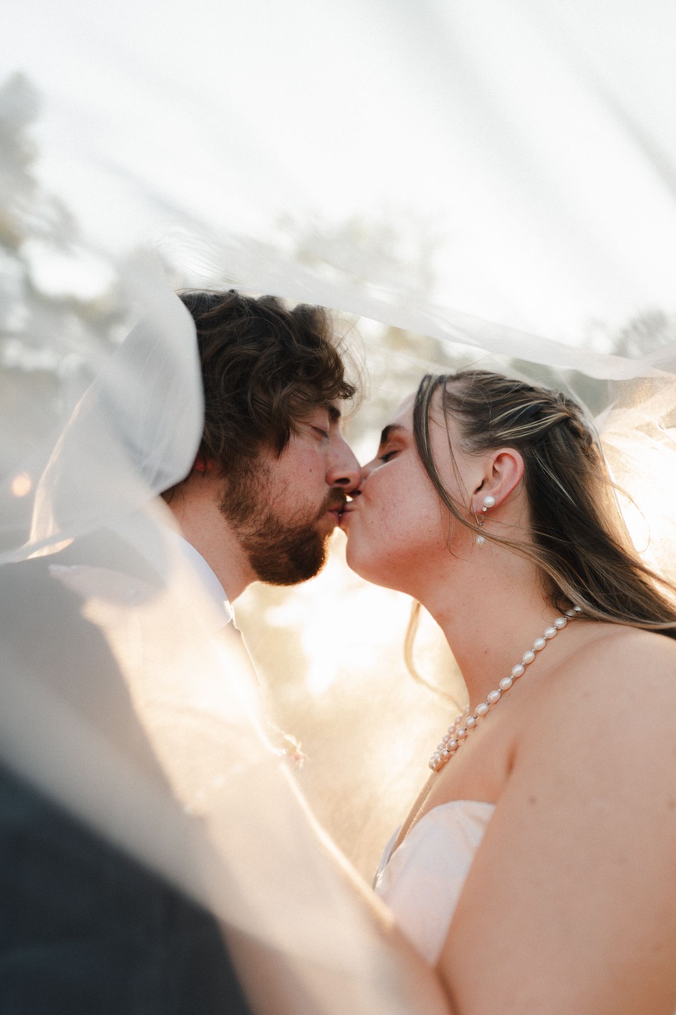 A bride and groom share a romantic kiss under a wedding veil with soft golden backlight.