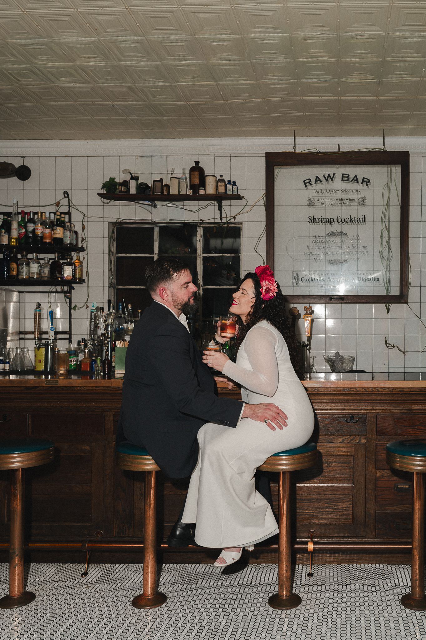 Bride and groom sitting at a vintage raw bar, sharing a cocktail and laughing together on their wedding day.