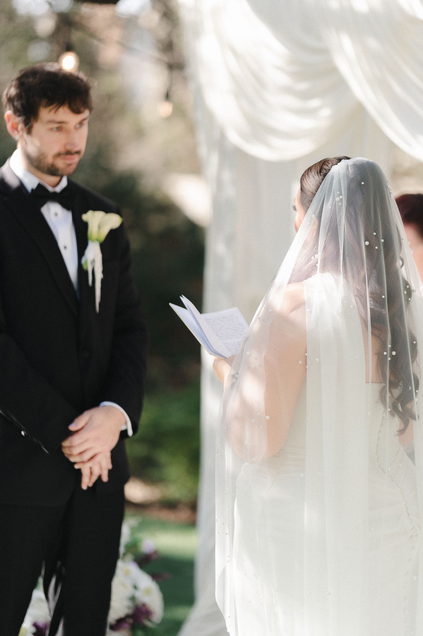 Bride reads vows to groom at outdoor wedding altar adorned with white draping and floral decor.