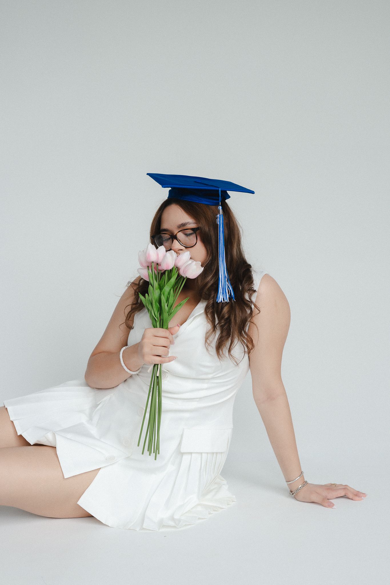 Young woman in white dress and blue graduation cap smelling pink tulips while sitting on white floor.