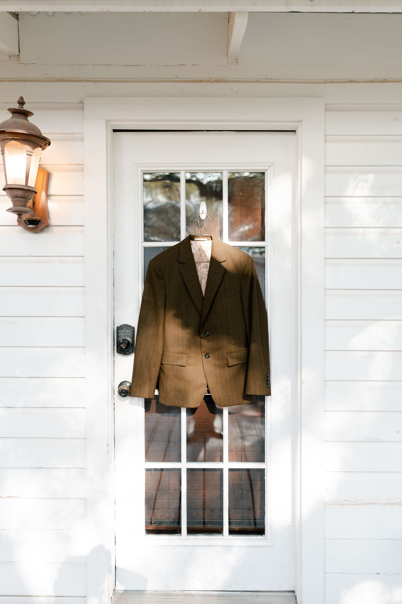 A brown suit jacket displayed on a hanger against a white door with glass panels and a vintage wall lantern.