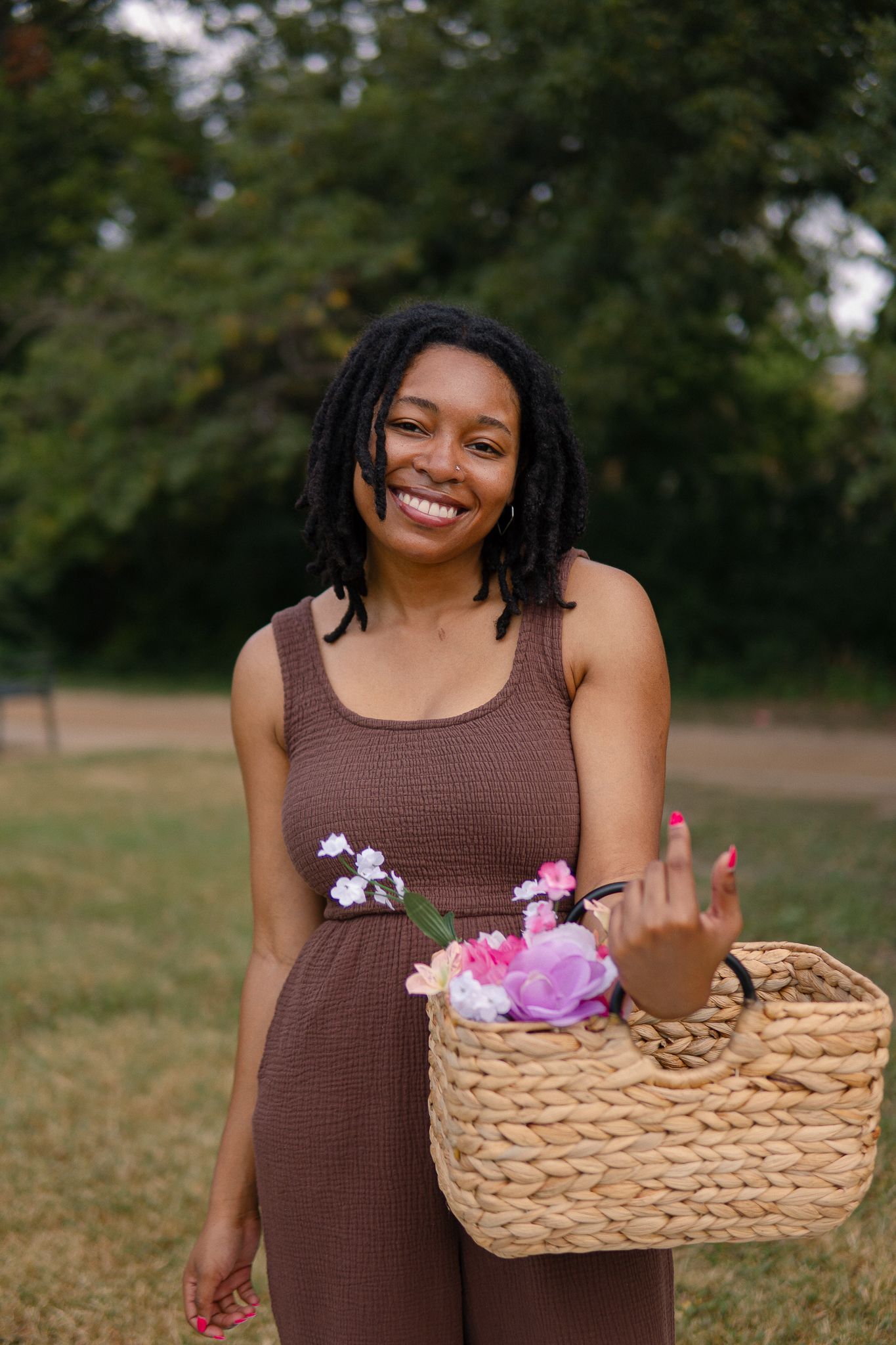 Person in brown dress holds woven basket with pink and purple flowers while standing in outdoor field.