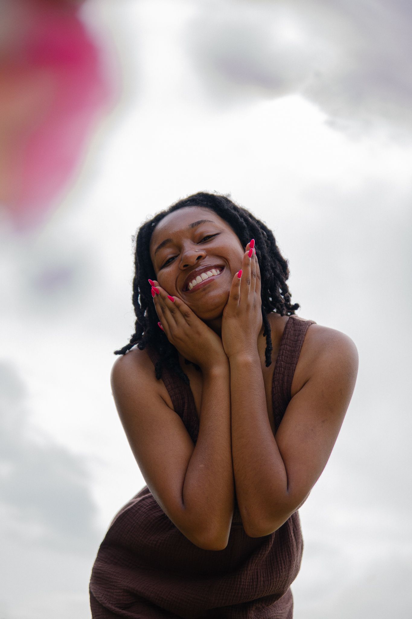 A joyful person wearing a brown top laughs while covering their face with their hands against a light background.
