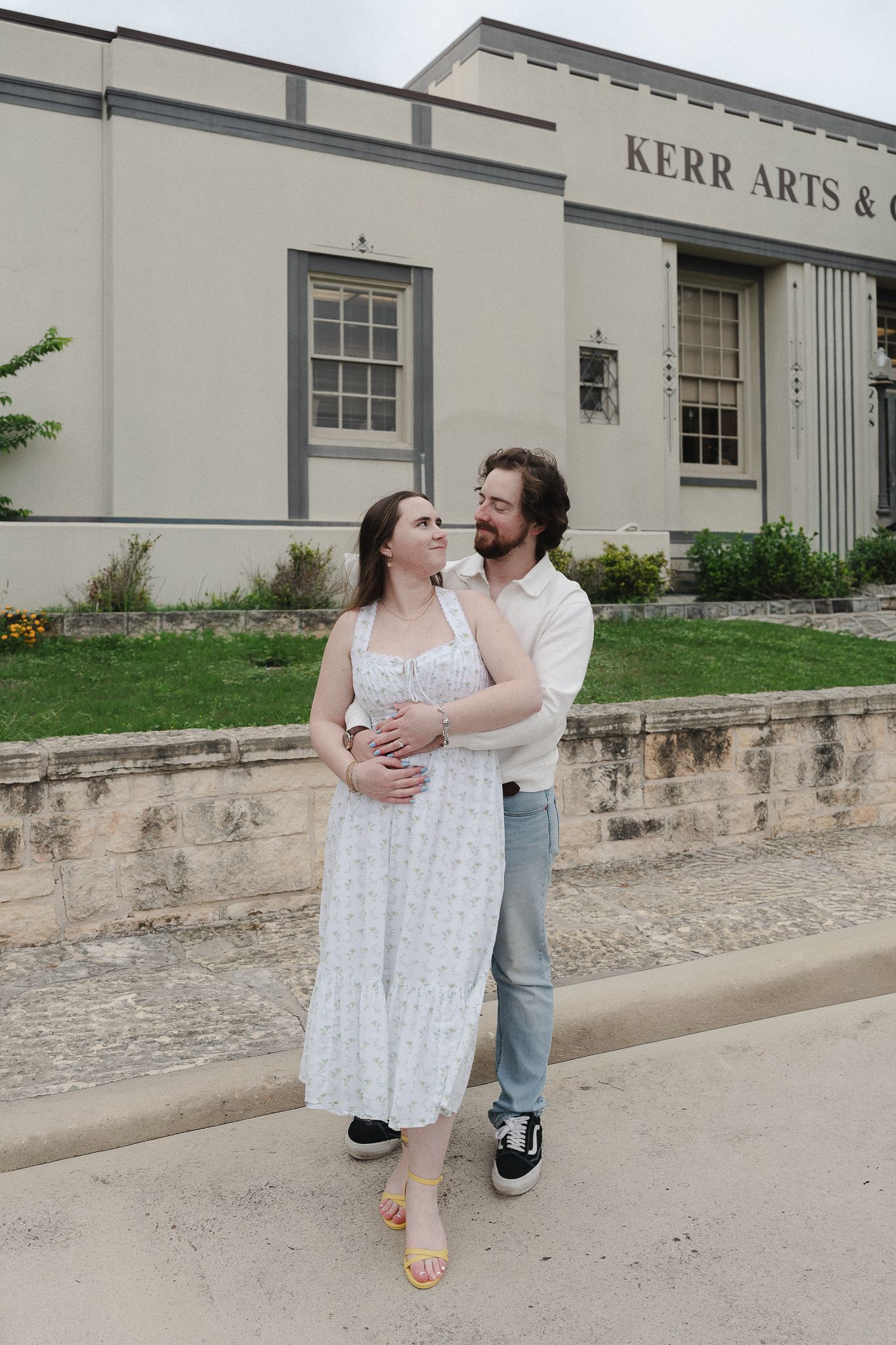 Couple embracing and smiling in front of Kerr Arts building, woman in white dress, man in white shirt and jeans.