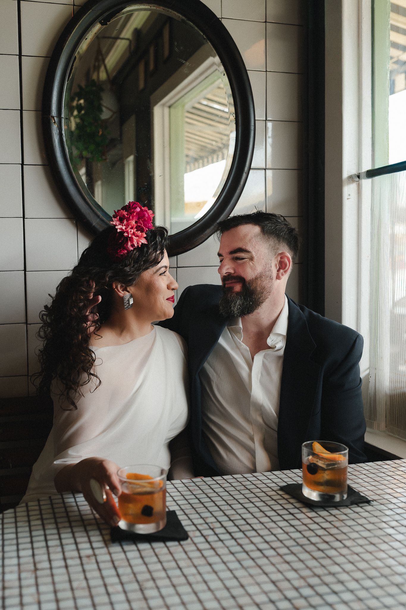Couple sharing cocktails at a tiled bar, woman with floral headpiece and man in suit gazing at each other romantically.
