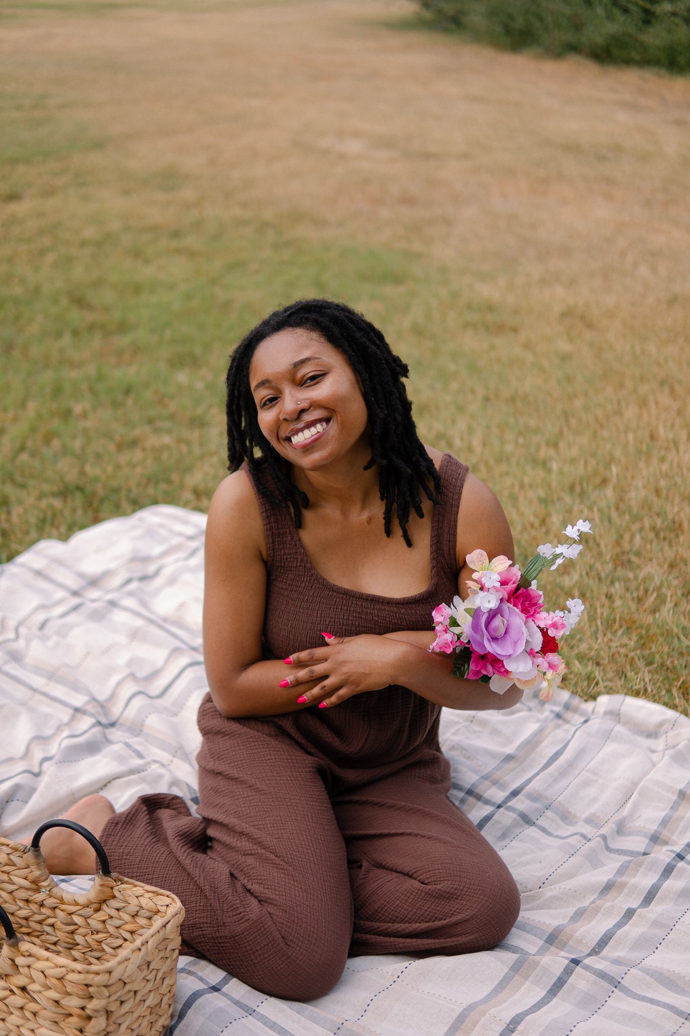 Someone sits on a picnic blanket in a brown jumpsuit holding pink flowers with a woven basket nearby.