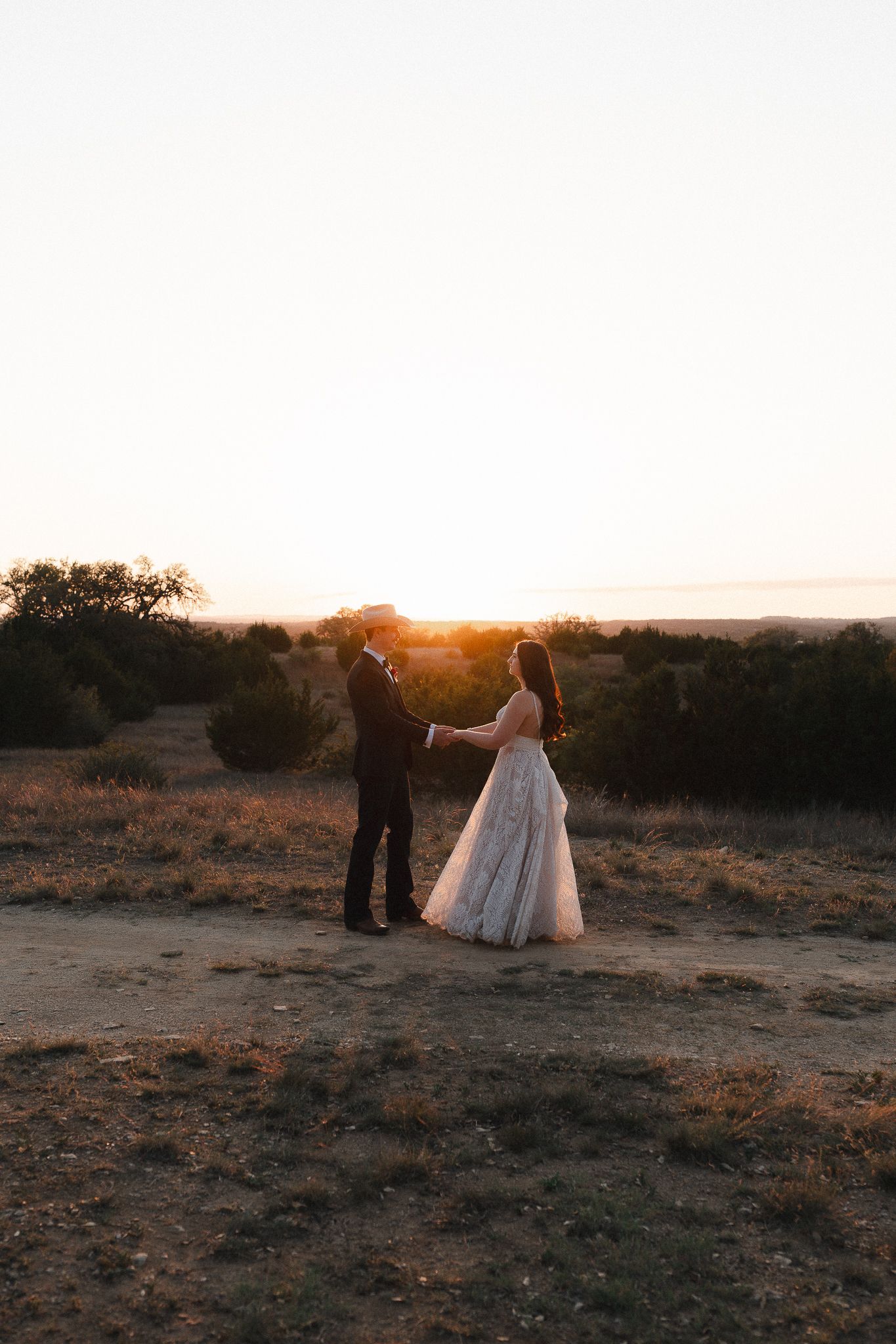 Bride and groom hold hands at golden sunset in rustic desert landscape, wearing wedding gown and dark suit.