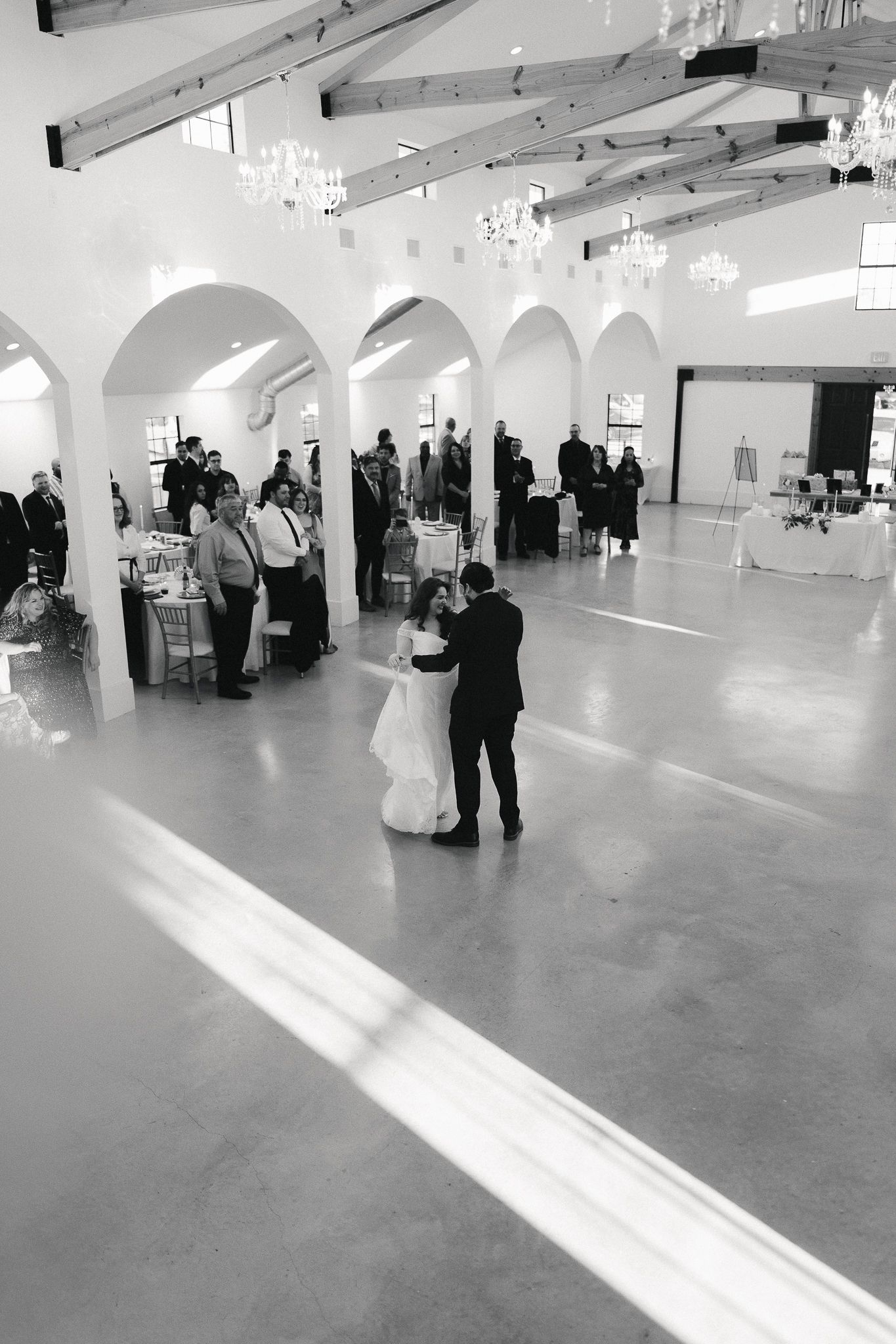 Black and white photo of a bride and groom sharing their first dance in an elegant white ballroom with arched columns.