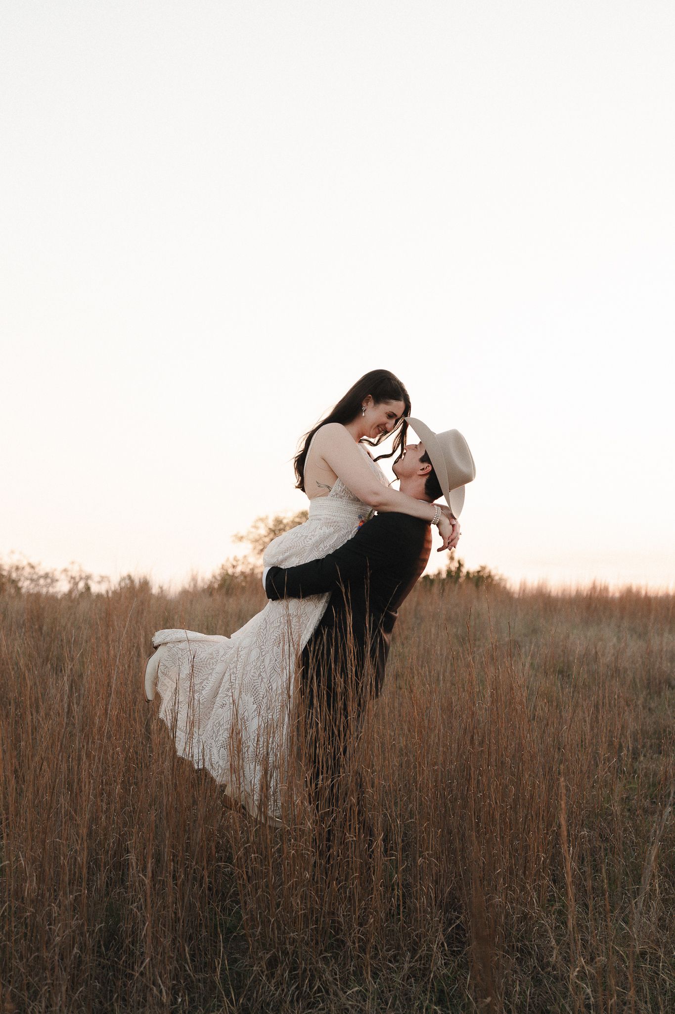 A groom lifts his bride in a sunlit field of tall golden grass, sharing a romantic moment at sunset.