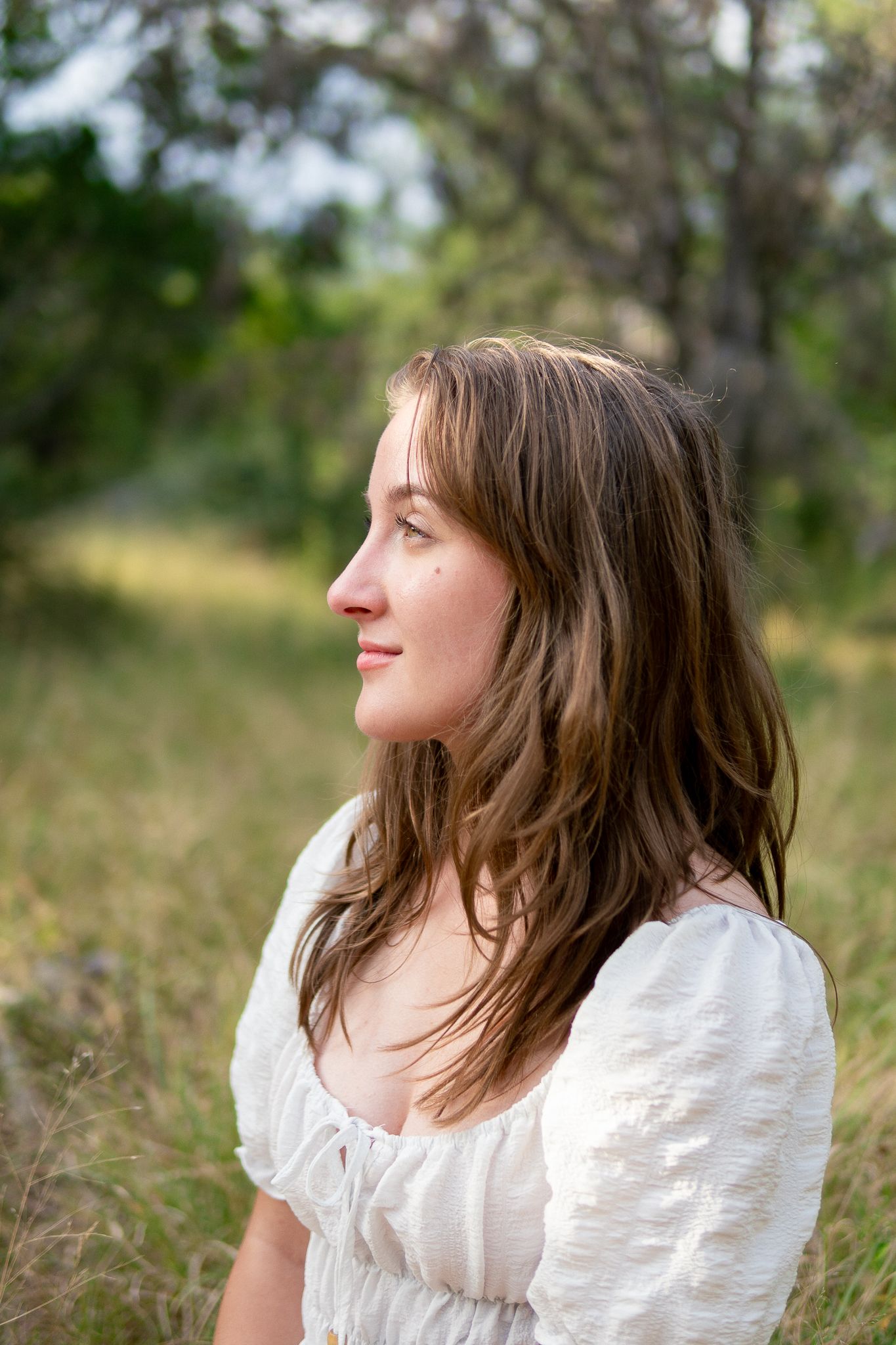 Profile view of someone in a white peasant blouse with long brown hair against a soft natural outdoor background.