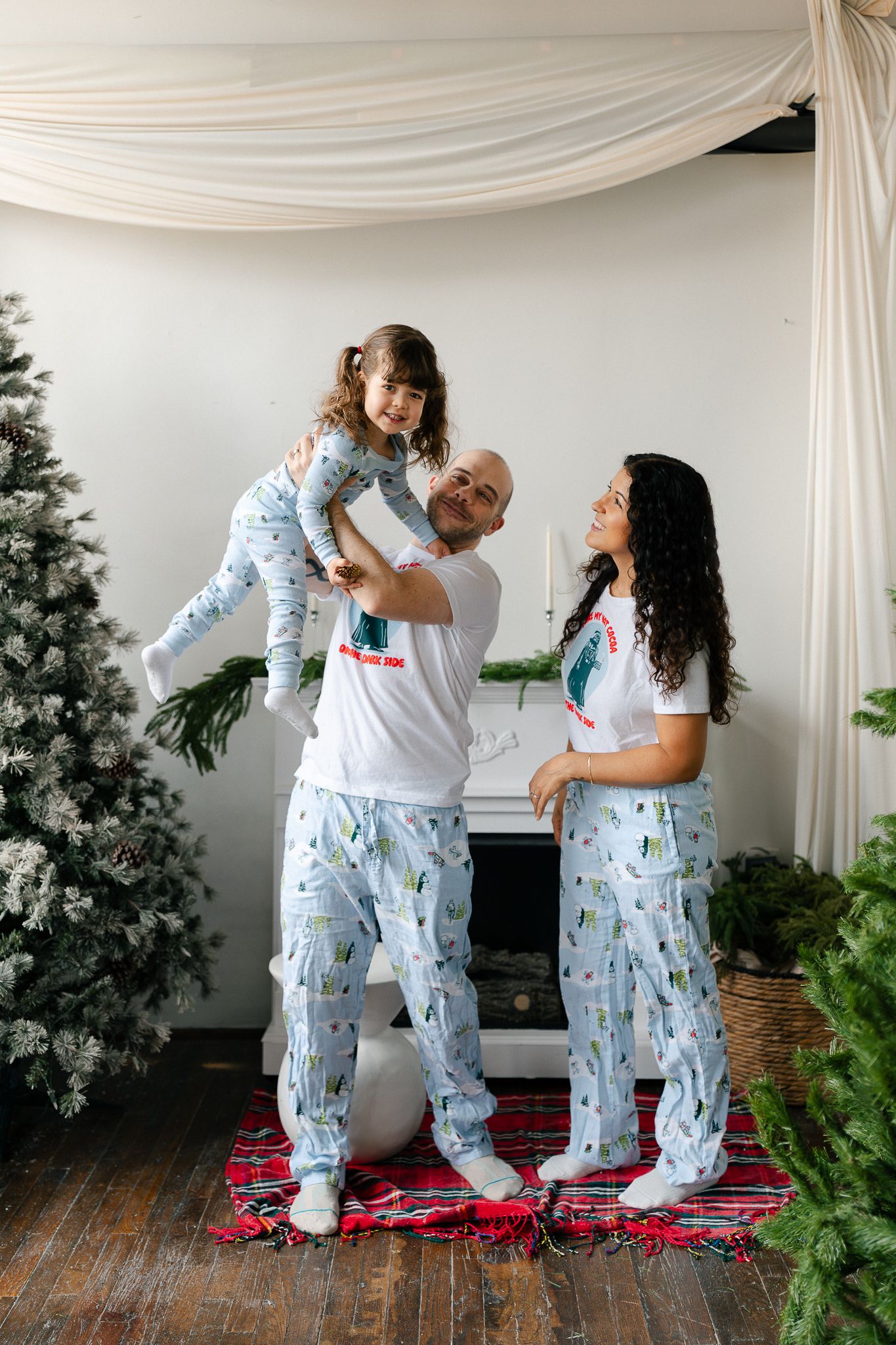 A family wearing matching winter-themed pajamas poses together in front of a Christmas tree and white draped backdrop.