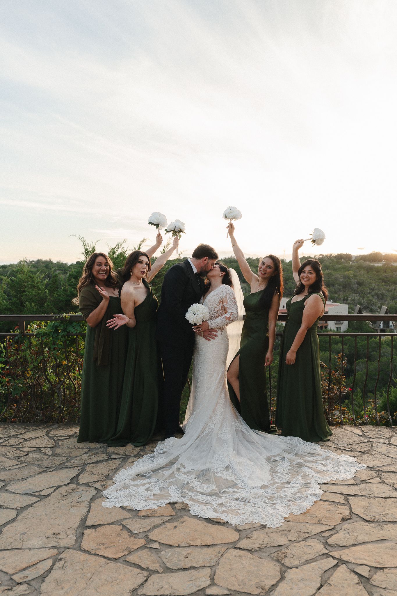Bride and groom kiss on a stone terrace at sunset while four bridesmaids in green dresses raise white bouquets.