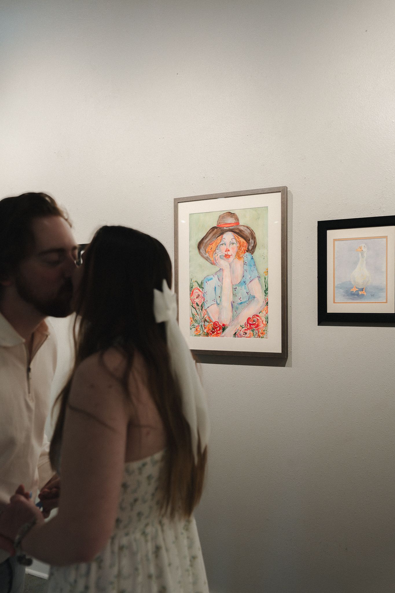 Couple sharing a kiss at an art gallery with colorful portrait painting of a red-haired woman in a hat on the wall.