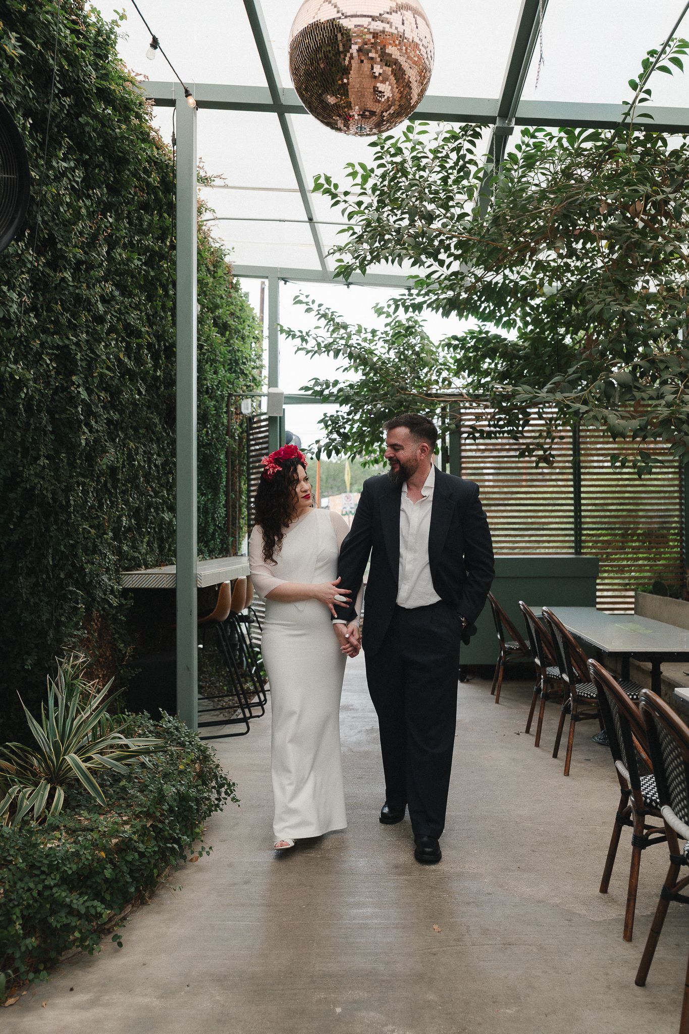 Bride and groom walk hand-in-hand through a lush greenhouse venue, bride wearing red floral headpiece and white gown.