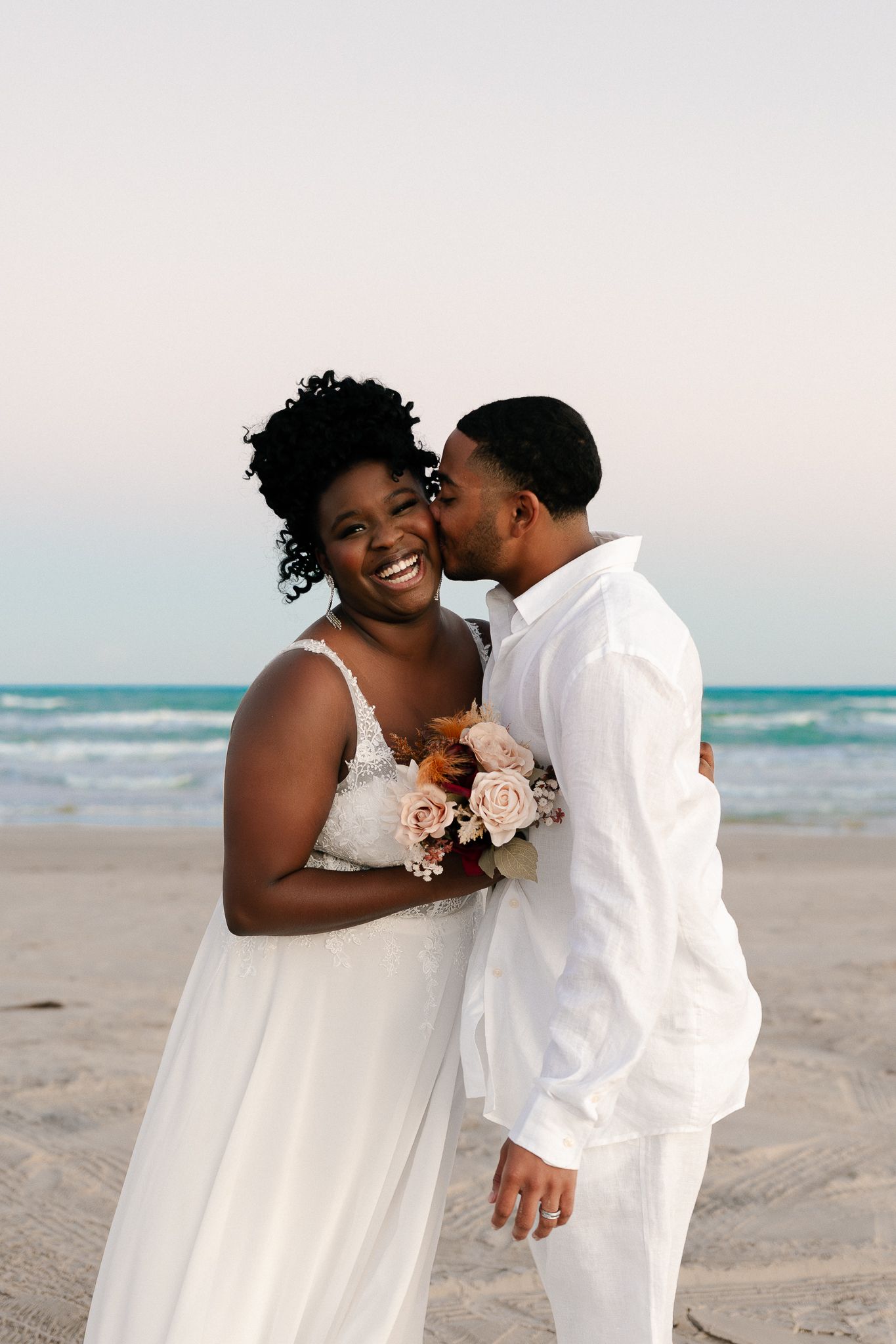 A romantic beach wedding photoshoot with a couple in white attire sharing intimate moments on sandy shores at sunset.