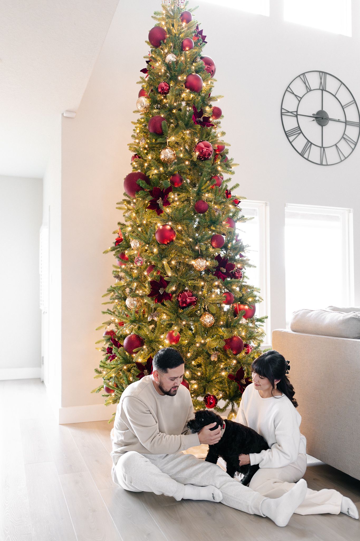 A beautifully decorated tall Christmas tree with burgundy and gold ornaments illuminates a bright modern living room.
