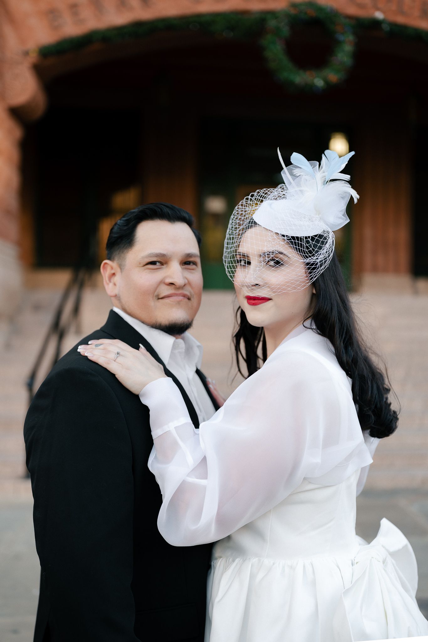 A couple poses for a wedding photo, with the bride wearing a vintage-inspired white dress and feathered fascinator headpiece.