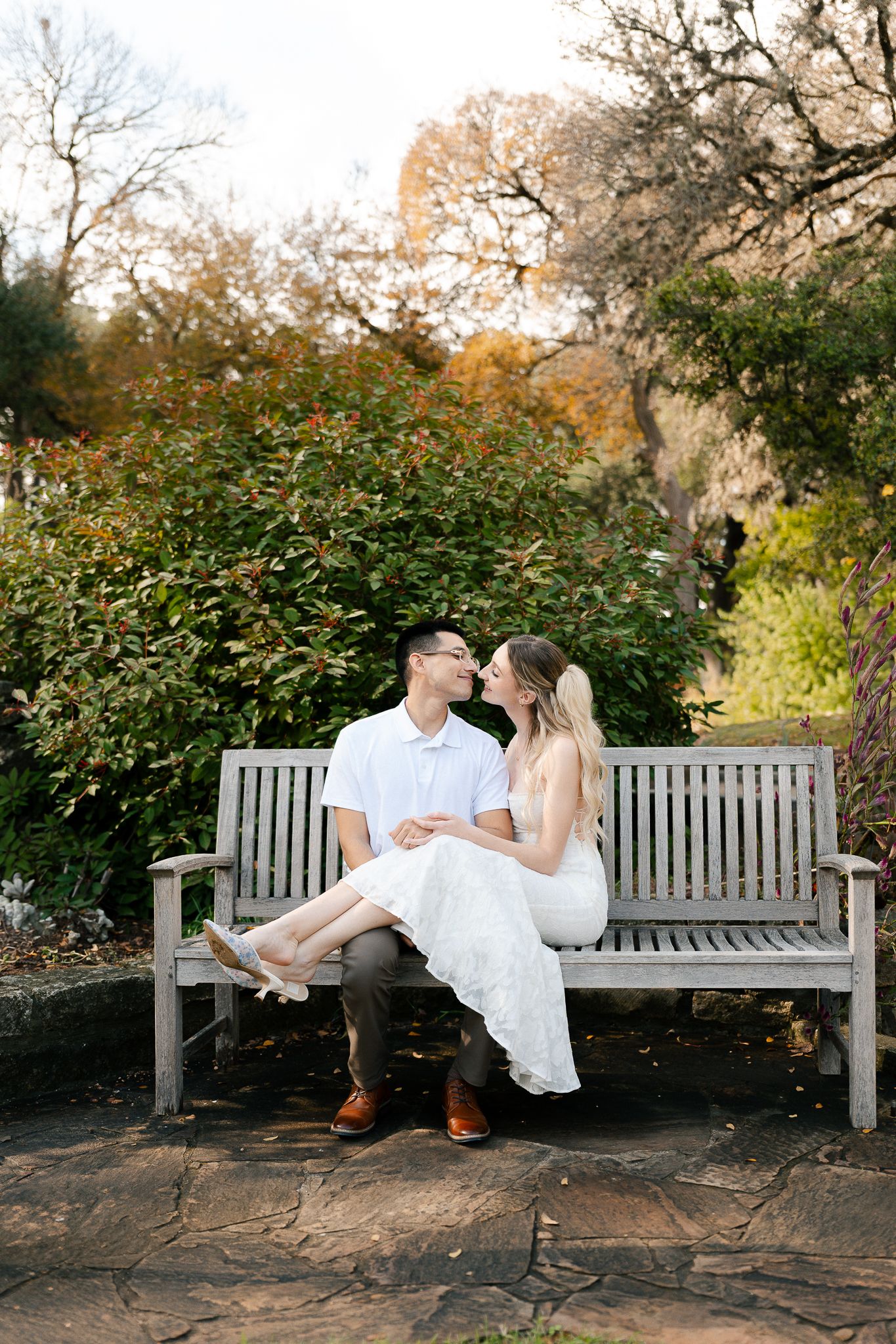 A couple in wedding attire sits together on a park bench in a romantic sequence of poses during an autumn day.