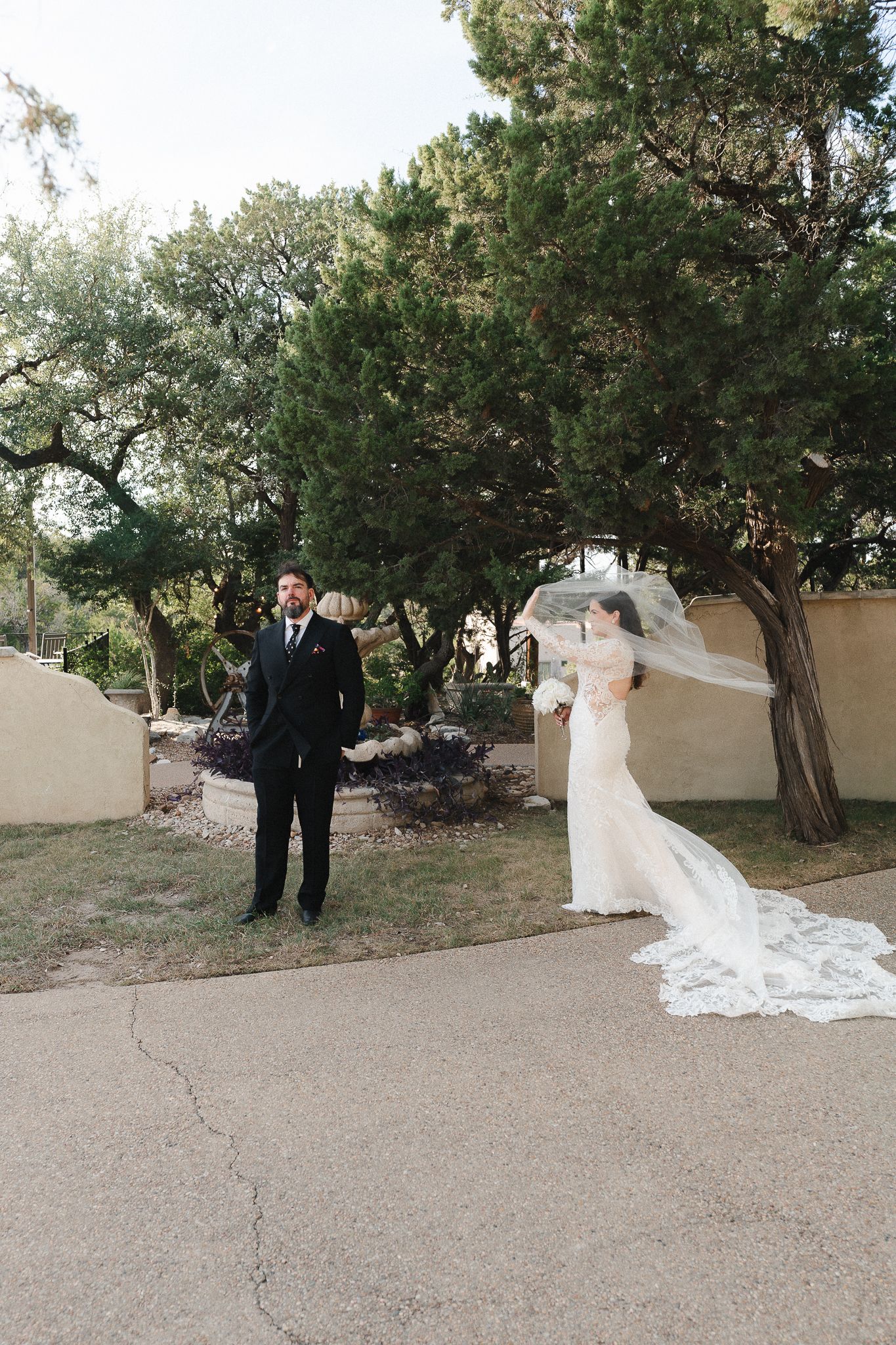 Bride in a lace wedding gown with a flowing veil stands behind a groom in a black suit during a first look under trees.