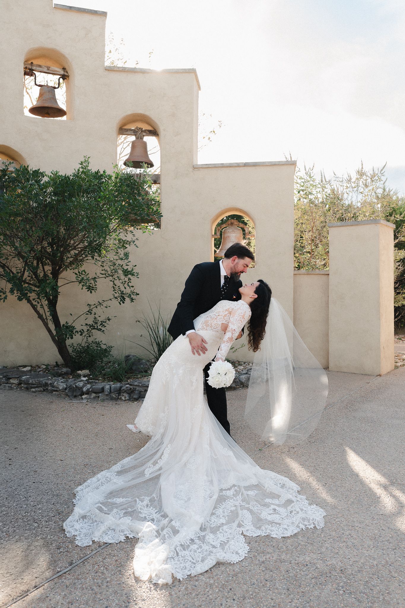 Groom dips bride in a white lace gown with a long veil outside a sunlit adobe-style chapel with bell arches.